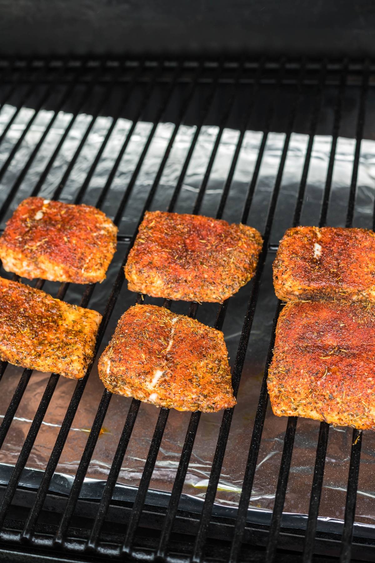 Six seasoned salmon fillets cooking on a grill with foil underneath the grates—a perfect smoked salmon recipe made in a pellet grill or Traeger for your next summer feast.