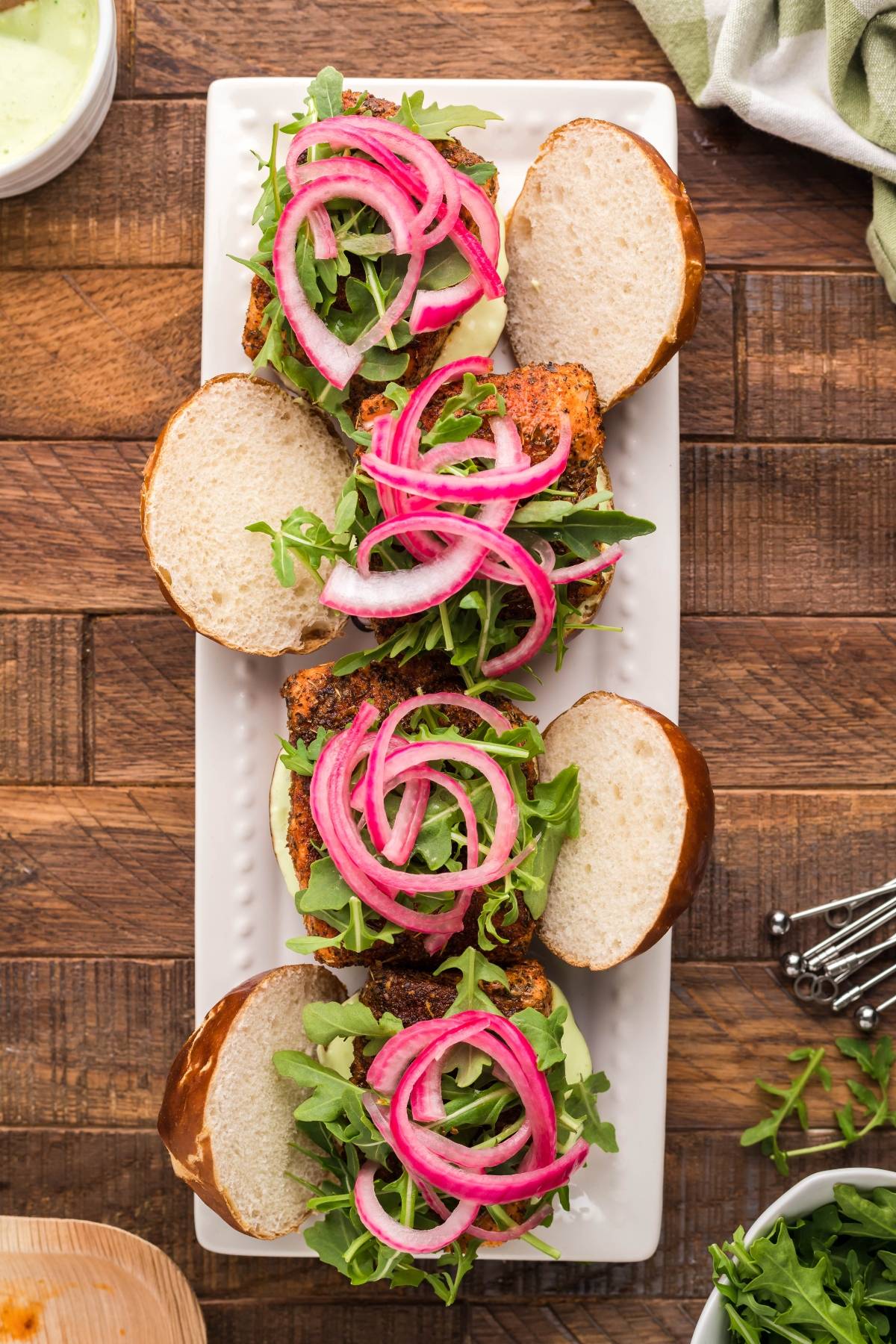 Three sandwiches with arugula, pickled red onions, and Wasabi Mayo on pretzel buns, arranged on a white rectangular plate—perfect for Summer BBQ salmon burgers.