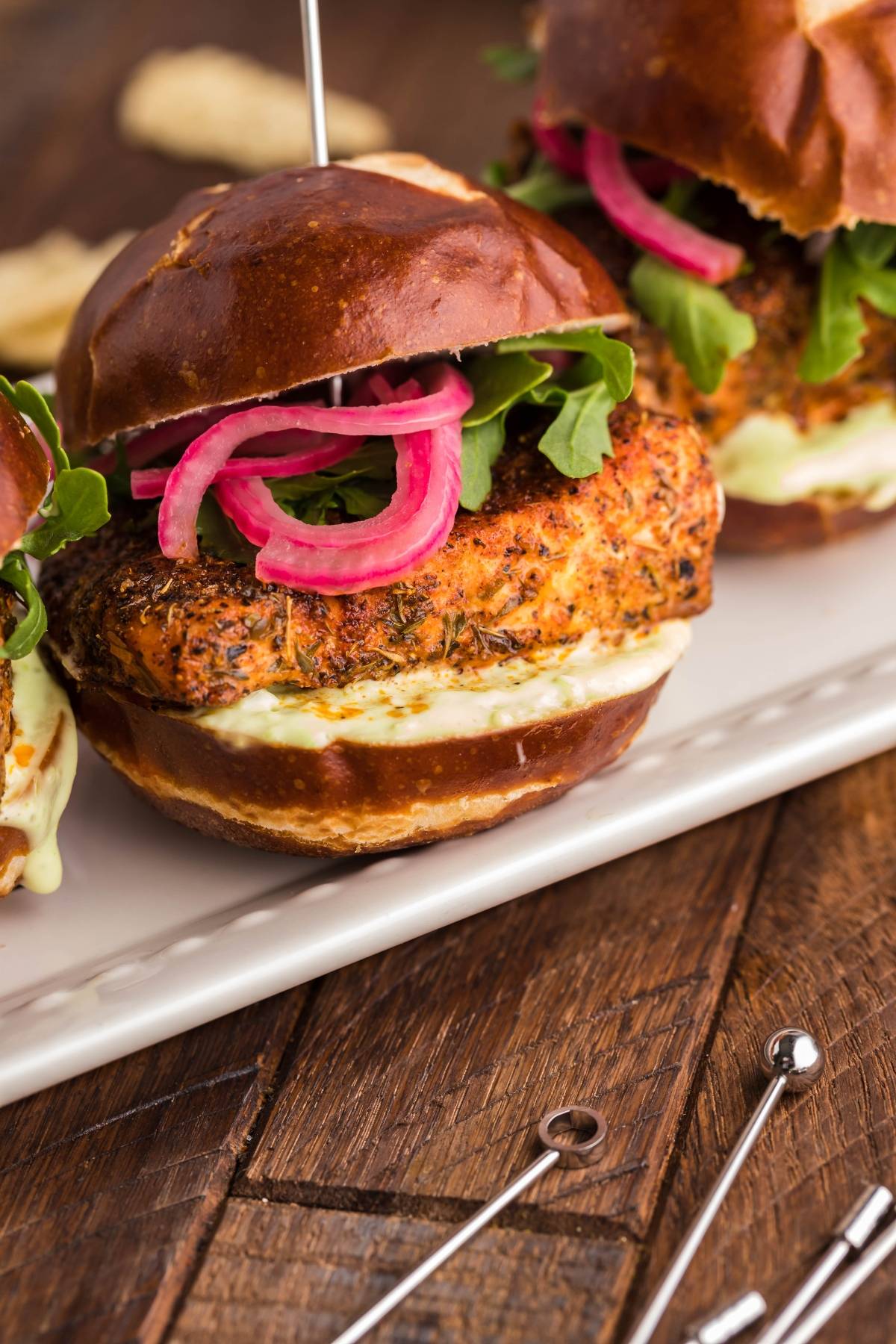 Chicken sandwich with lettuce, pickled onions, and sauce on a pretzel bun, served on a white platter—perfect alongside Smoked Salmon Sliders made in a pellet grill or Traeger for an irresistible meal.