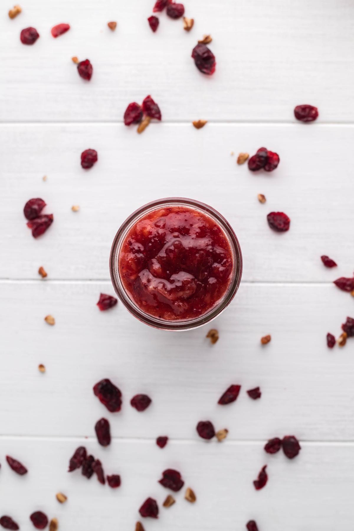 A jar of cranberry sauce with bourbon and pineapple sits on a white table, surrounded by scattered dried cranberries and nuts.