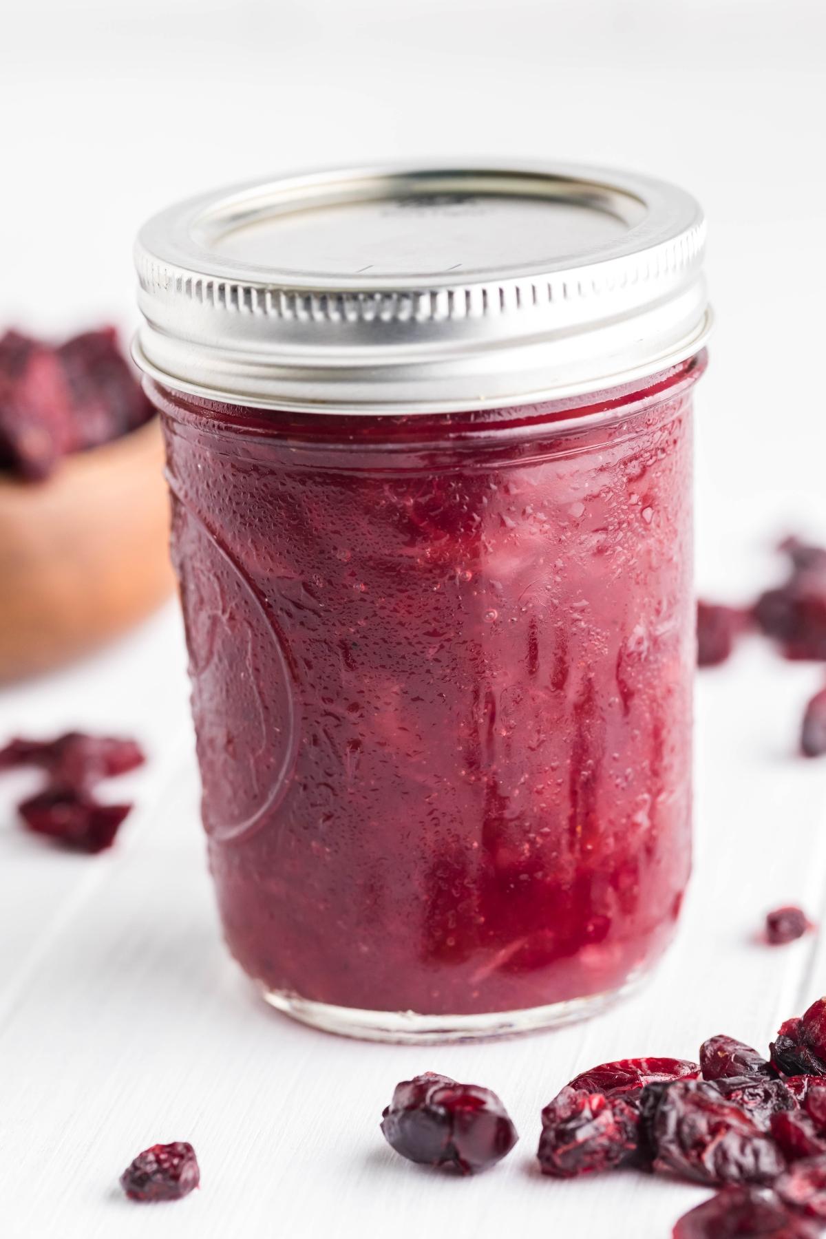 A glass jar filled with Spiked Cranberry Sauce, surrounded by dried cranberries on a white surface, makes this cranberry bourbon relish perfect for any festive table.