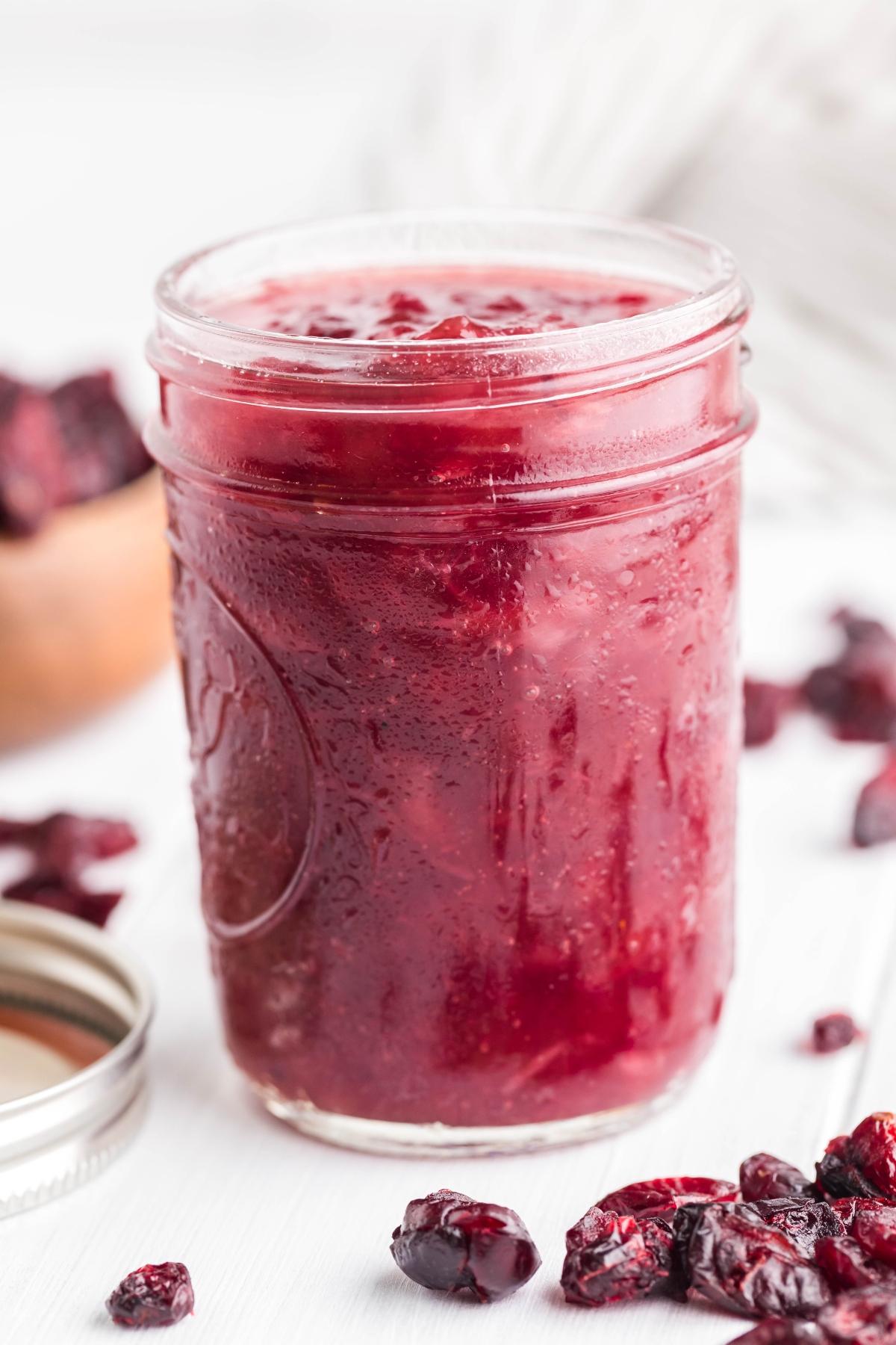 A glass jar filled with Spiked Cranberry Sauce, with dried cranberries scattered around it, adds a festive twist to your table.