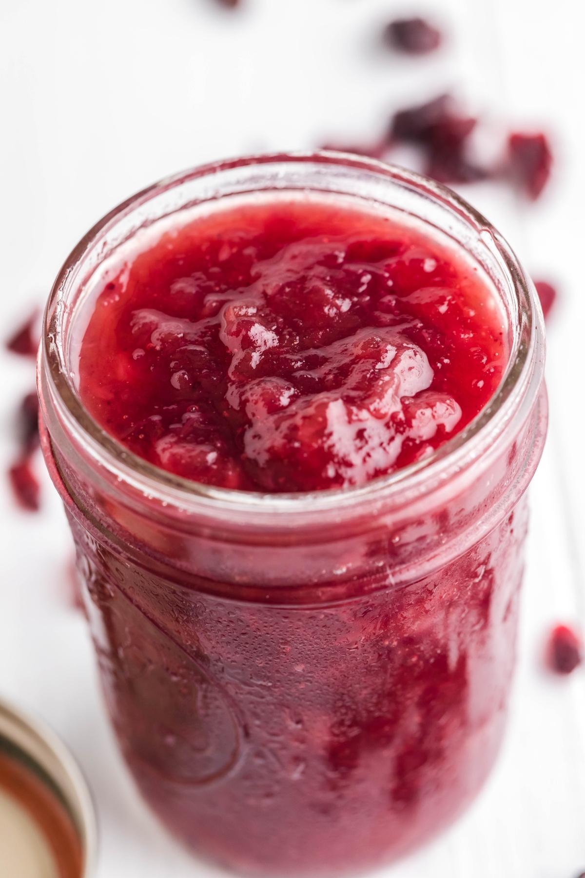Close-up of a glass jar filled with bright red, chunky spiked cranberry sauce on a white surface.