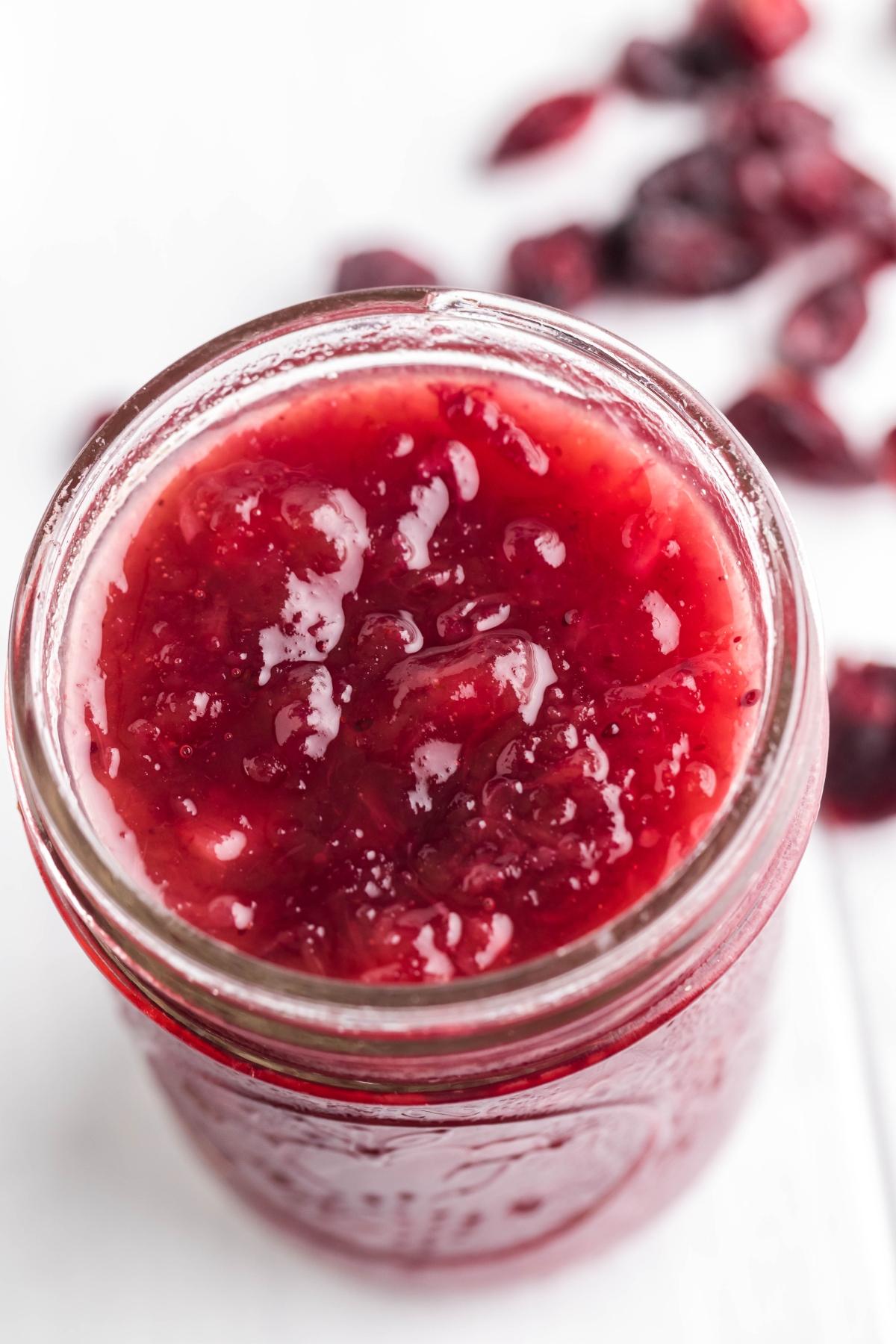 A jar of bright red spiked cranberry sauce sits amid scattered cranberries in the background, adding a festive twist to your holiday table.