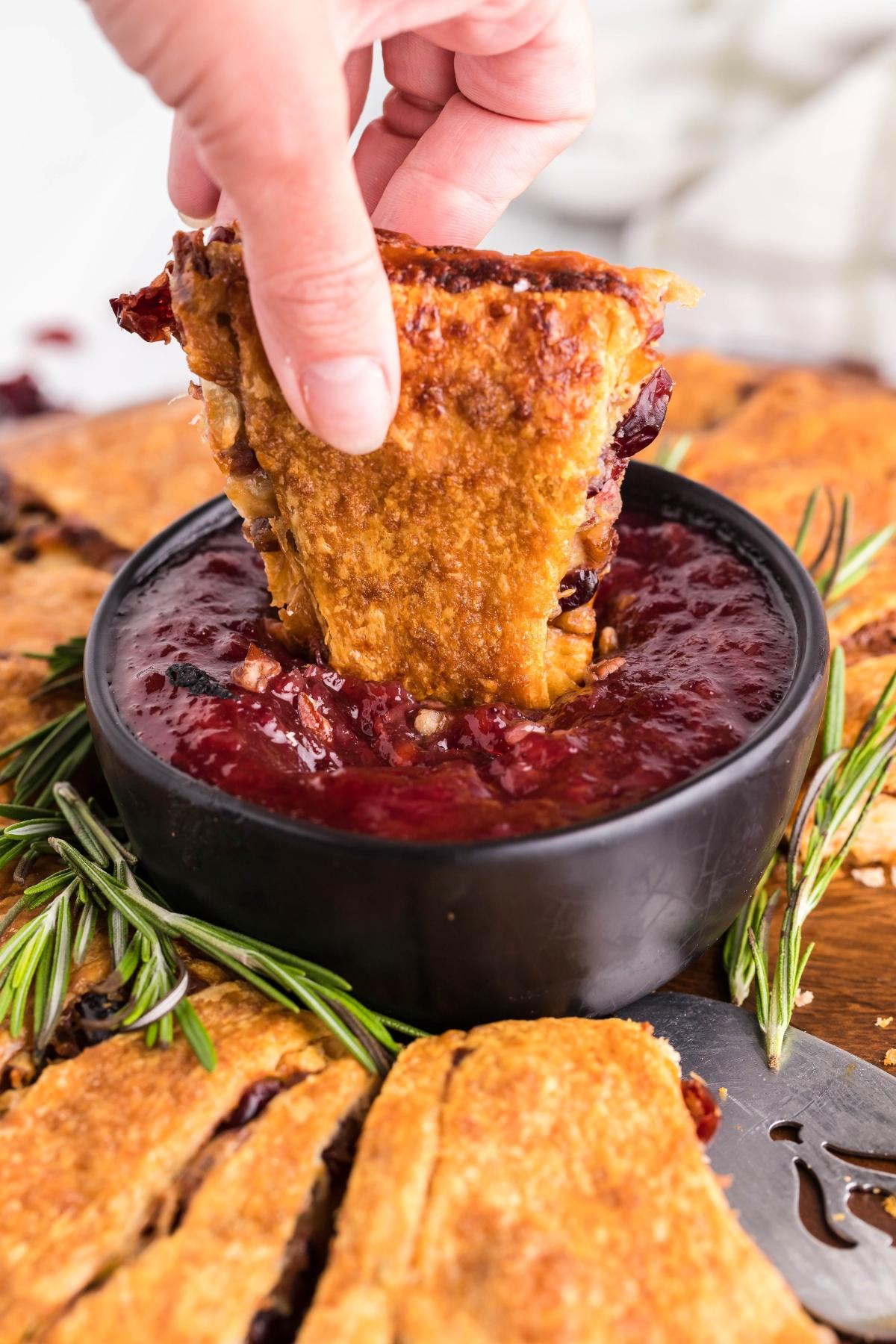 A hand dips a slice of pie into a bowl of spiked cranberry sauce, garnished with fresh rosemary.