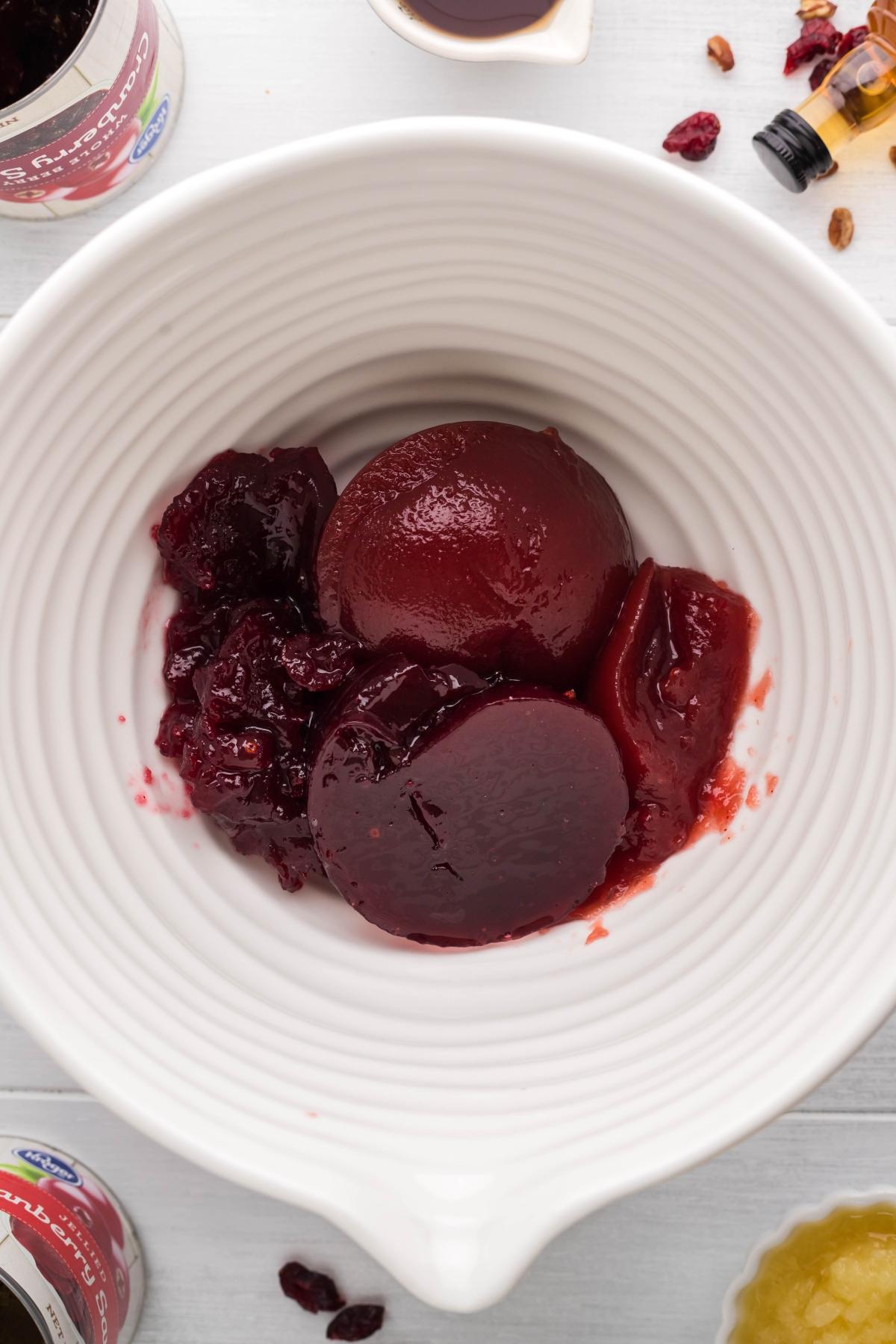 Canned cranberry sauce in a white bowl on a light surface, with sauce and ingredients nearby—perfect for creating a spiked cranberry sauce recipe or transforming into a tasty cranberry bourbon relish.