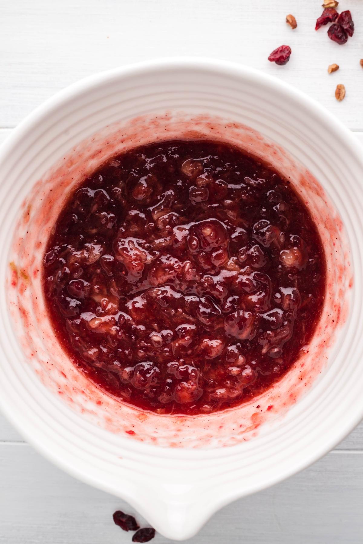 A white bowl filled with chunky cranberry sauce on a light surface, featuring a cranberry bourbon relish, with a few cranberries nearby.