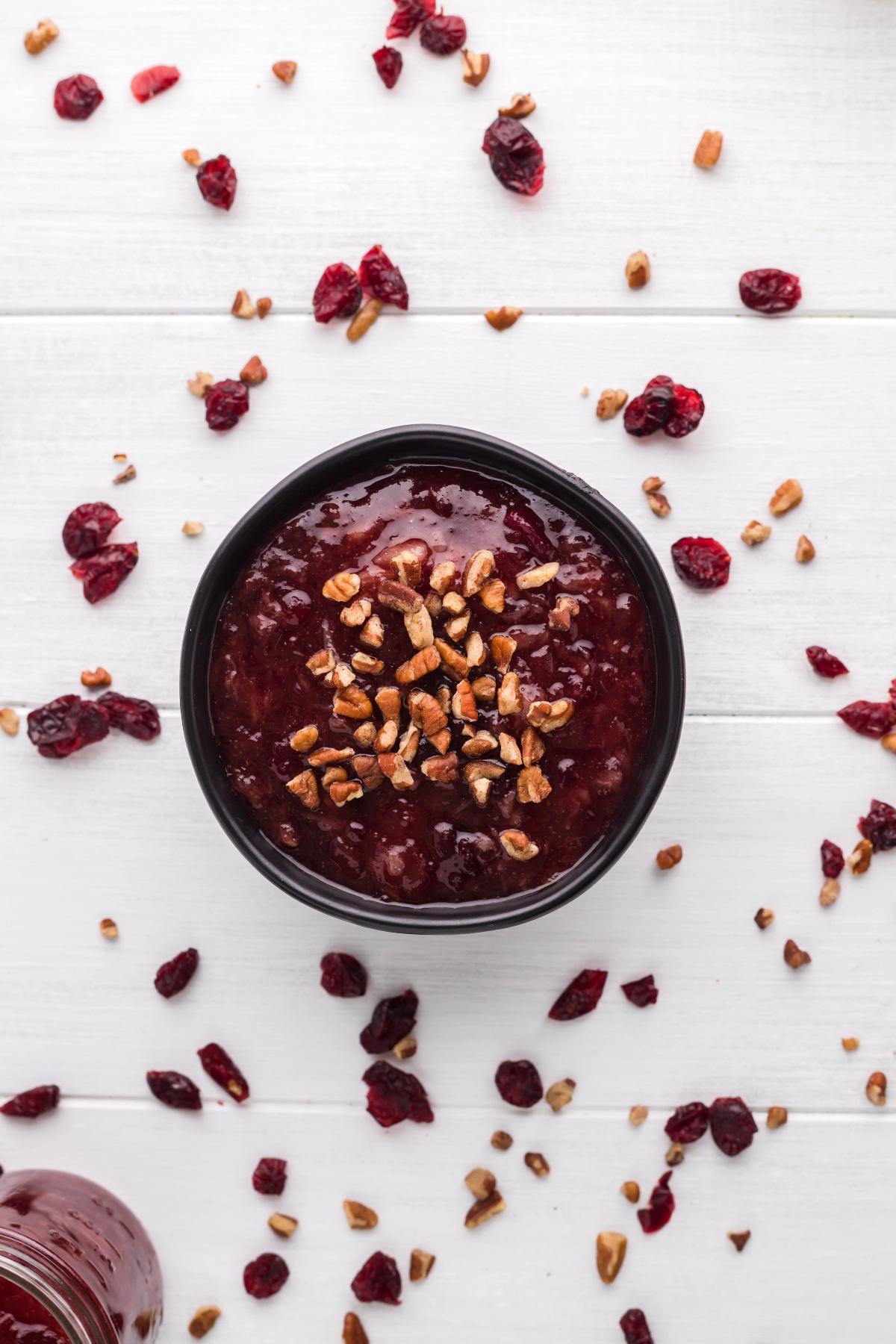 A bowl of Spiked Cranberry Sauce topped with chopped nuts, surrounded by dried cranberries on a white surface.