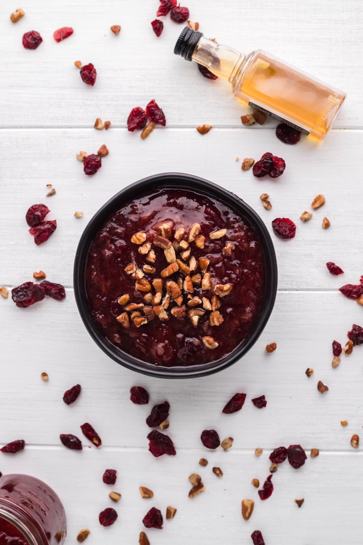 Bowl of Spiked Cranberry Sauce topped with chopped pecans, surrounded by dried cranberries, nuts, and a small bottle—perfect for trying a cranberry sauce with bourbon and pineapple twist.