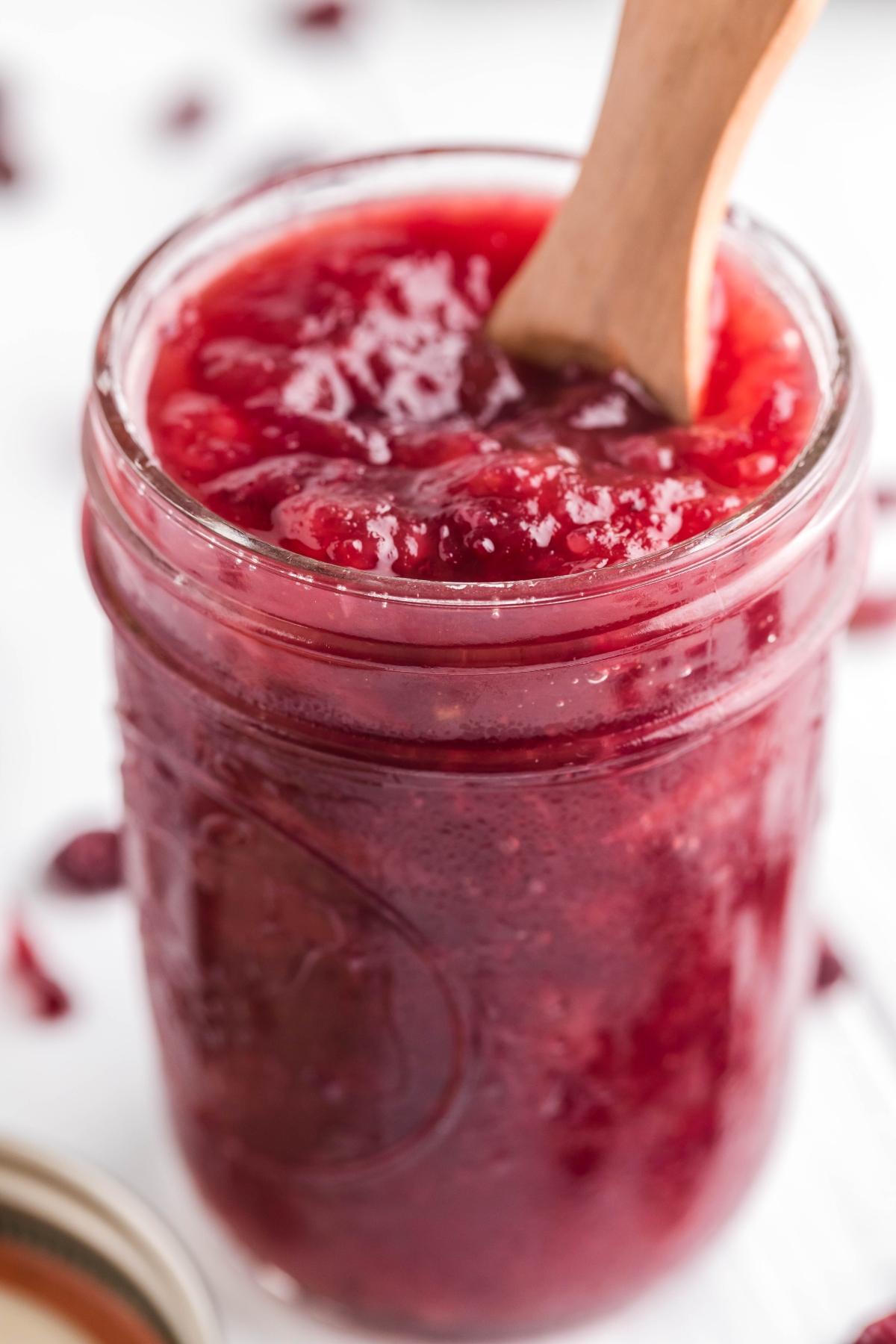 A jar of chunky Spiked Cranberry Sauce with a wooden spoon inside.