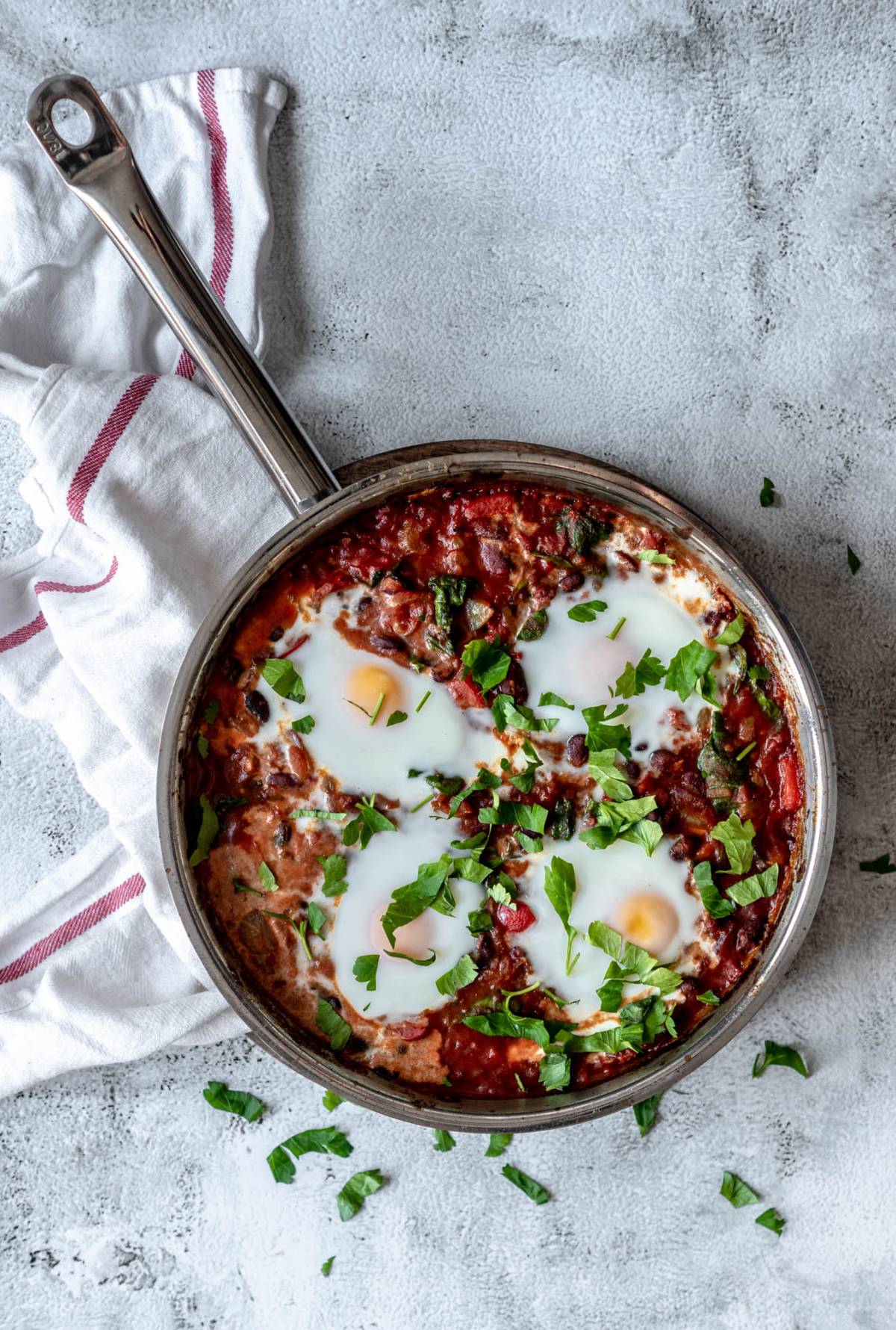A skillet of savoury breakfast recipe shakshuka with poached eggs, tomato sauce, and fresh herbs on a light surface with a towel nearby—a quick healthy meal to start your day.