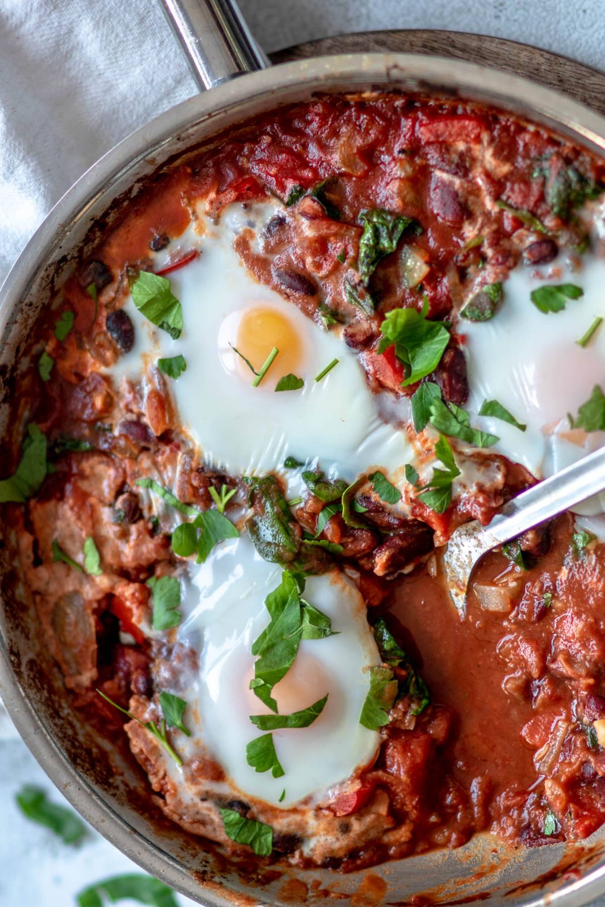 A pan of shakshuka featuring eggs poached in a tomato sauce with greens and fresh parsley on top—a savoury breakfast recipe that's also a quick healthy meal.