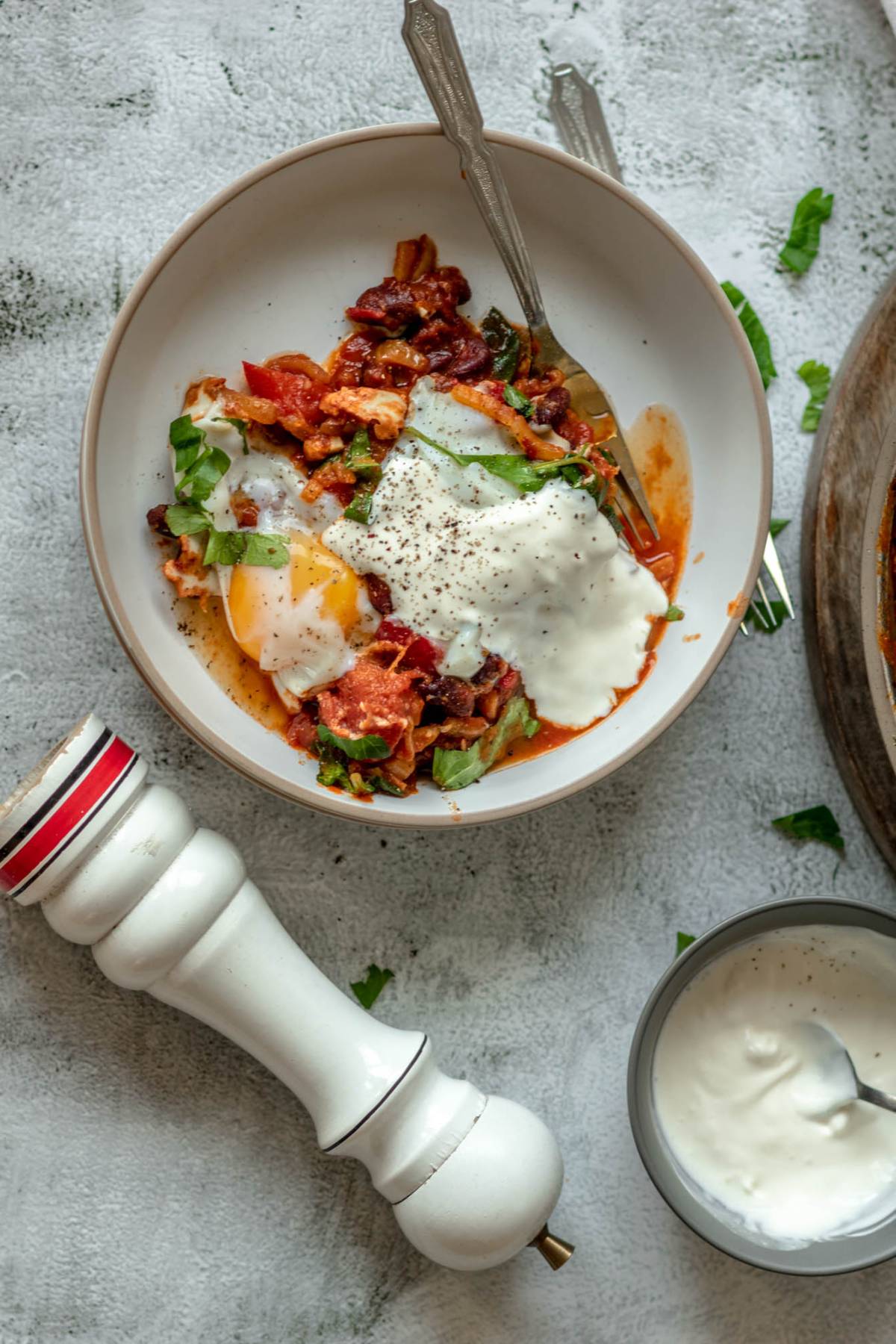 A bowl of shakshuka with eggs poached in a tomato sauce, topped with yogurt and herbs, sits beside a pepper mill and small bowl of yogurt—a quick healthy meal perfect for any time of day.
