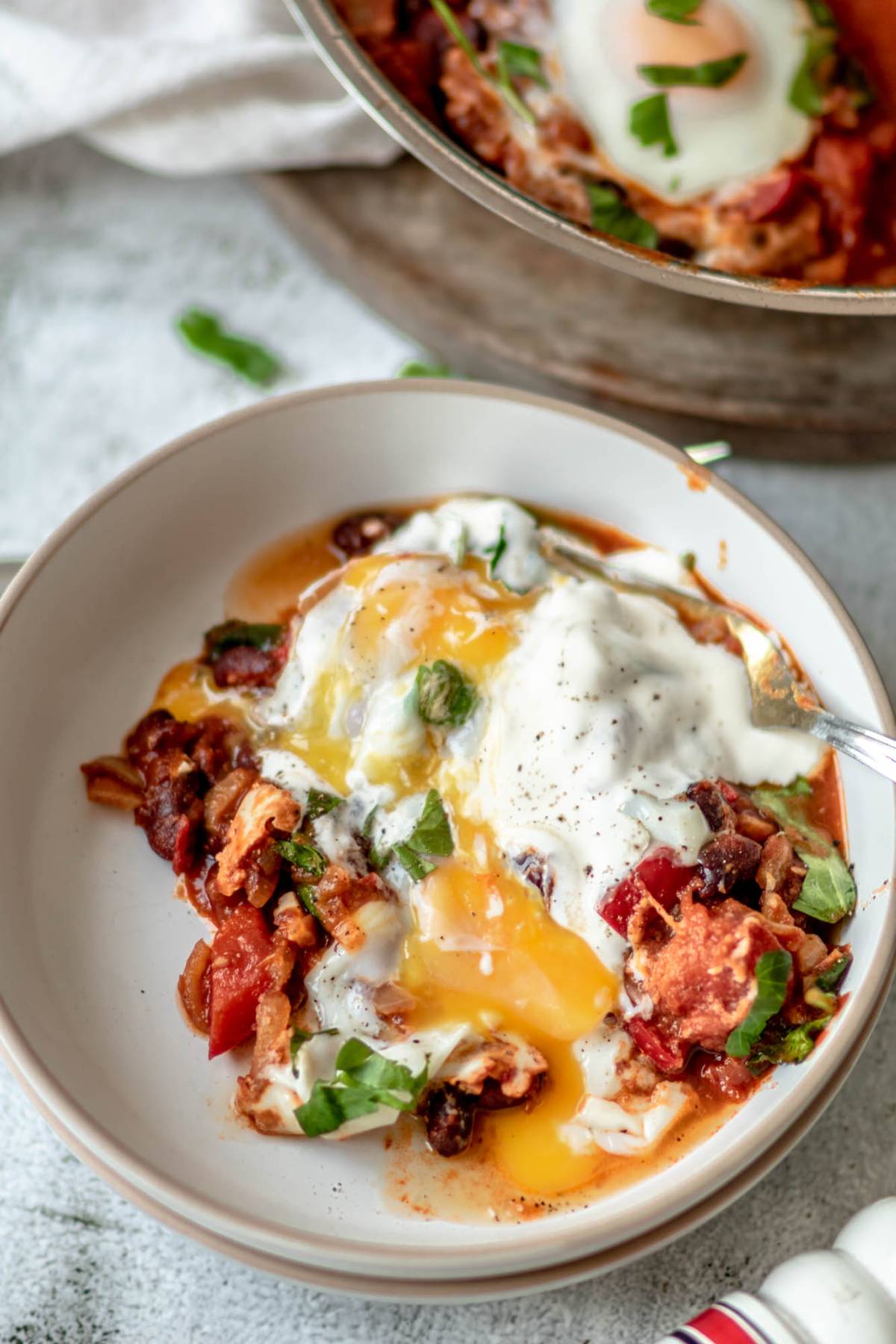 A plate of shakshuka with eggs poached in a tomato sauce, runny yolk, fresh herbs, and a dollop of yogurt on top—an easy one pot meal with spinach for added flavor.