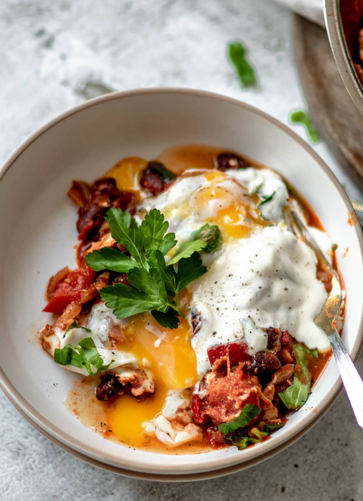 A bowl of savoury breakfast shakshuka with eggs poached in a tomato sauce, topped with herbs and yogurt garnish.