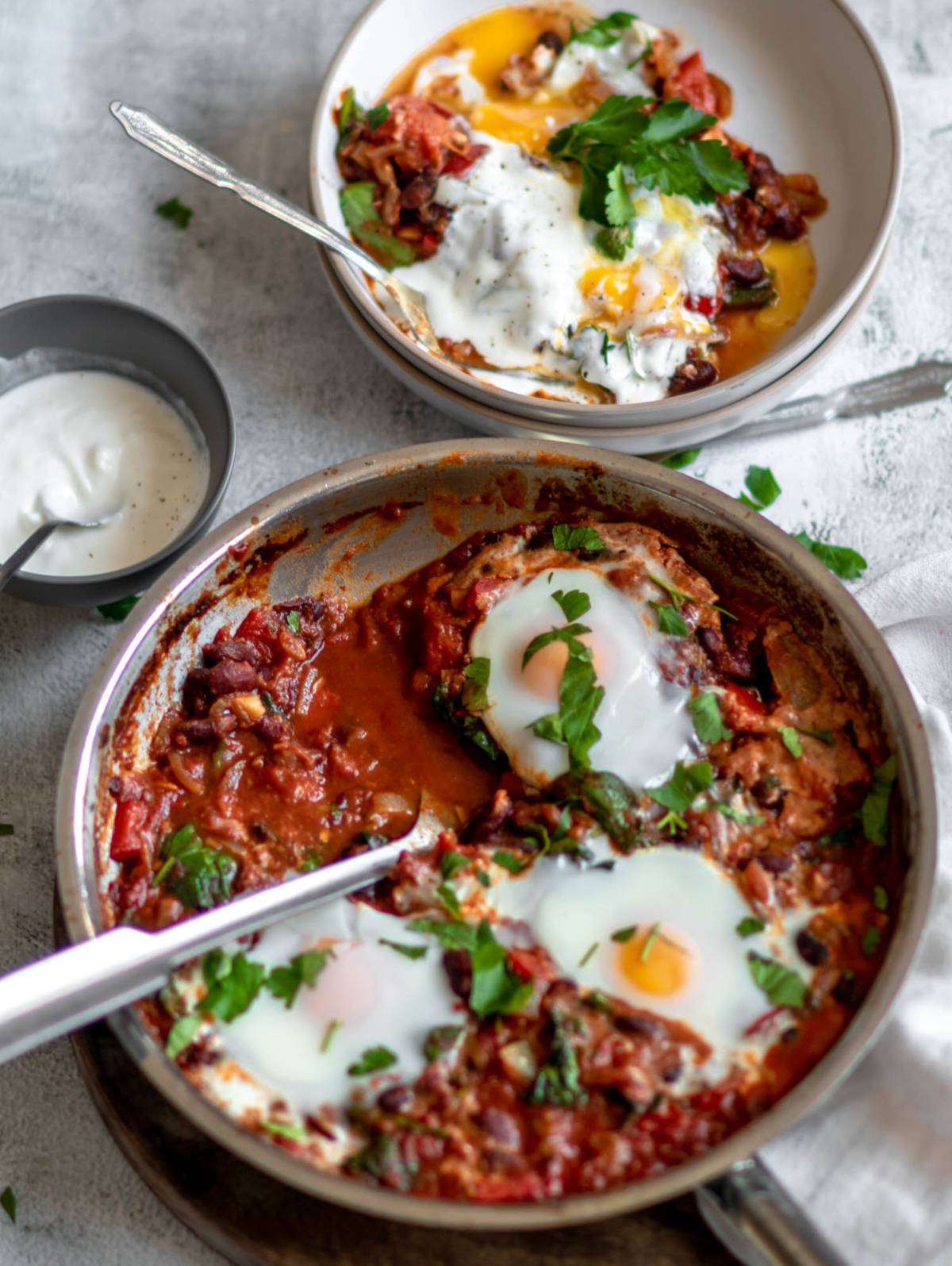 A pan of shakshuka with eggs poached in a tomato sauce, garnished with herbs, and a bowl of yogurt on the side—a quick healthy meal perfect for any time.