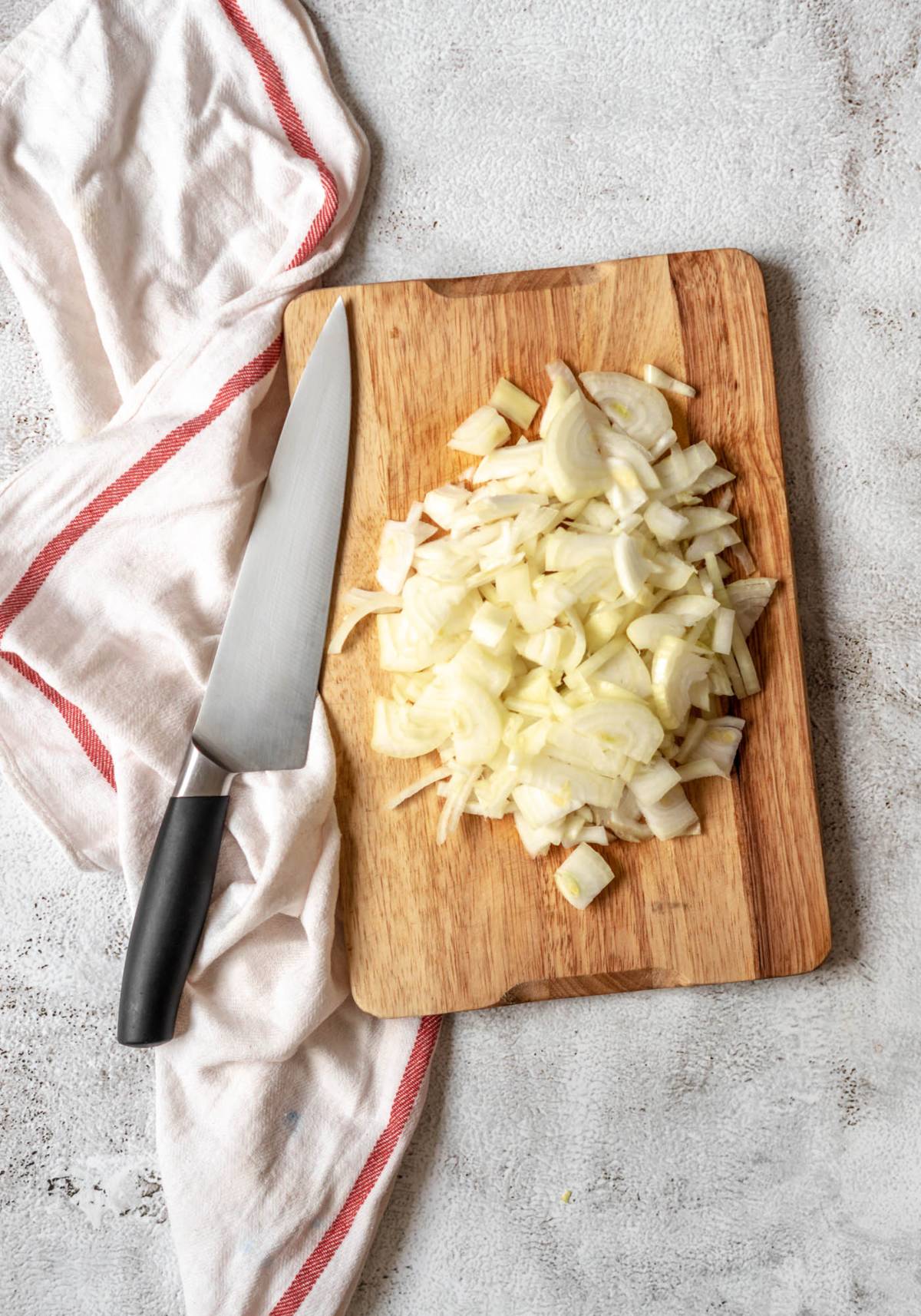 Chopped onions on a wooden cutting board with a knife and a white towel with a red stripe beside it—perfect prep for quick healthy meal recipes like spinach and red bean shakshuka.