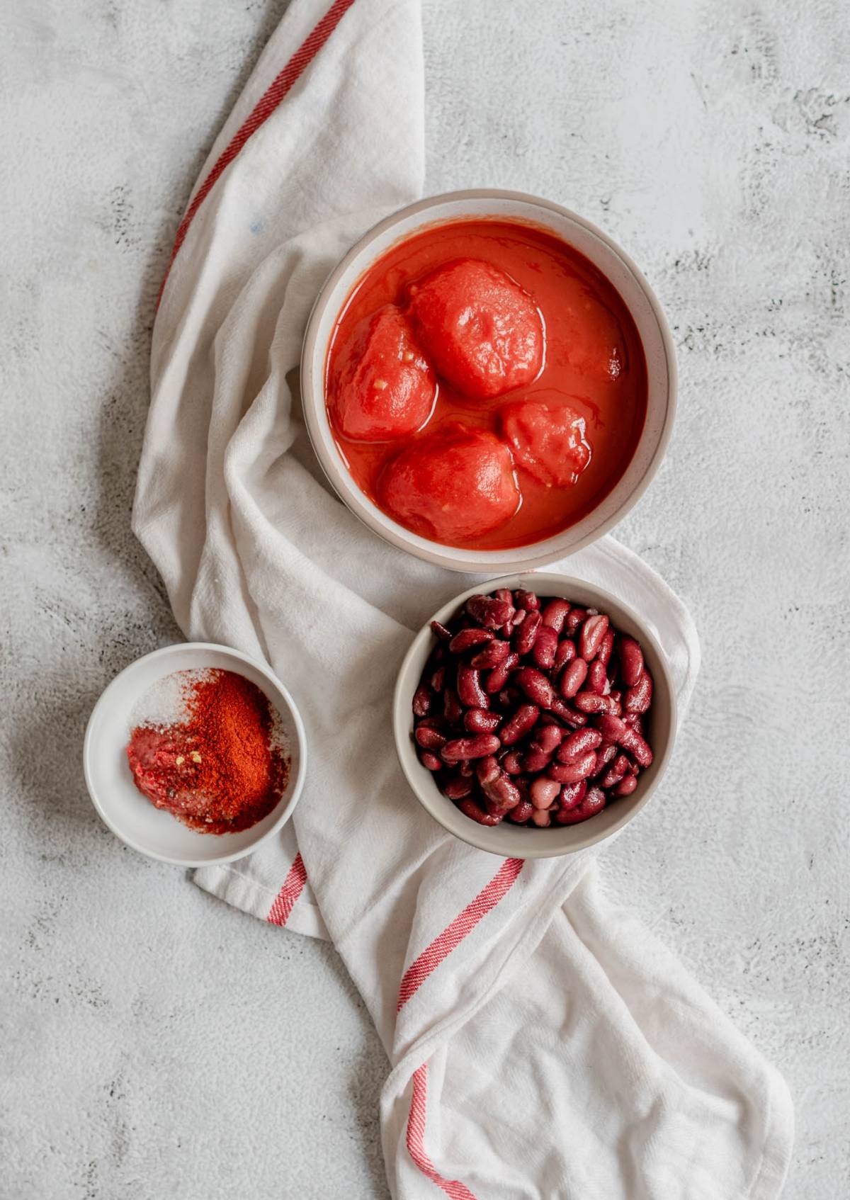 Three bowls with canned tomatoes, kidney beans, and spices on a white cloth with red stripes—perfect for a savoury breakfast recipe like eggs poached in a tomato sauce.