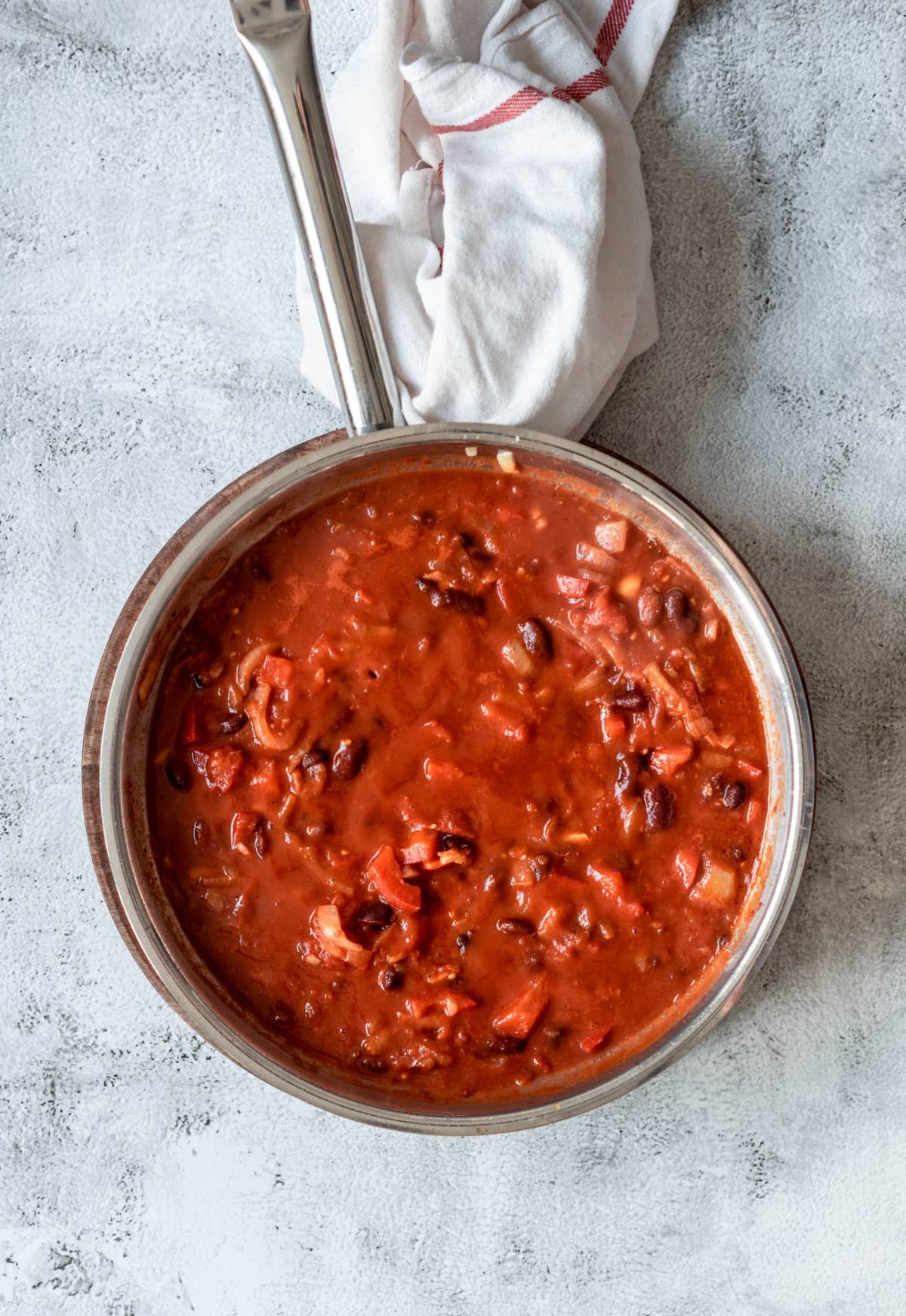 A saucepan filled with tomato and bean chili, a delicious one pot meal with spinach, sits on a gray surface with a white towel beside it.