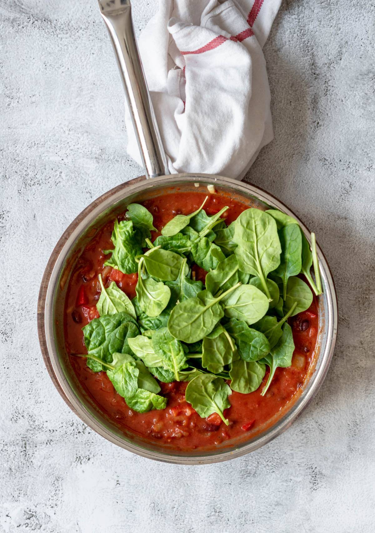 A pan of tomato sauce topped with fresh spinach leaves, perfect for a quick healthy meal, sits on a light gray surface with a white cloth nearby.