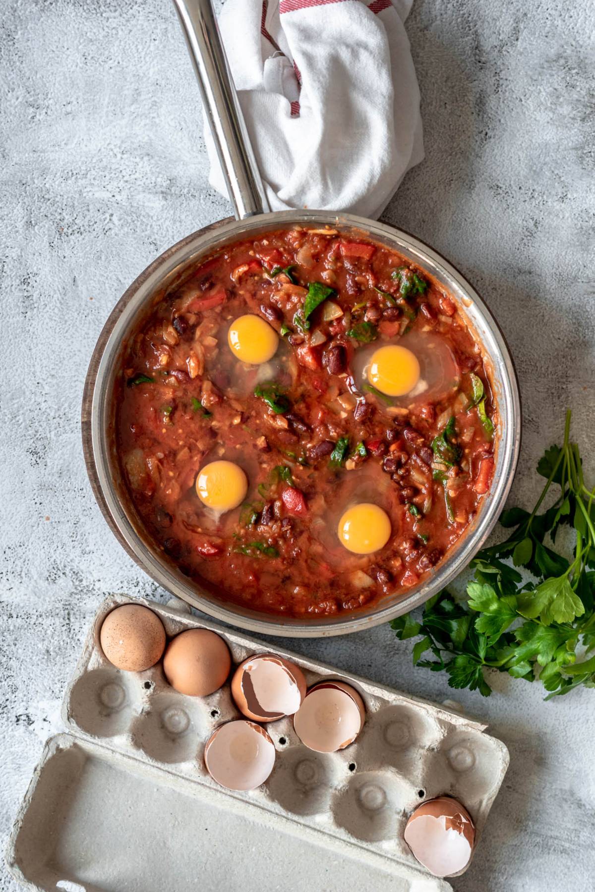 A skillet of eggs poached in a tomato sauce with fresh parsley sits on a gray surface, eggshells nearby—perfect as a quick healthy meal.