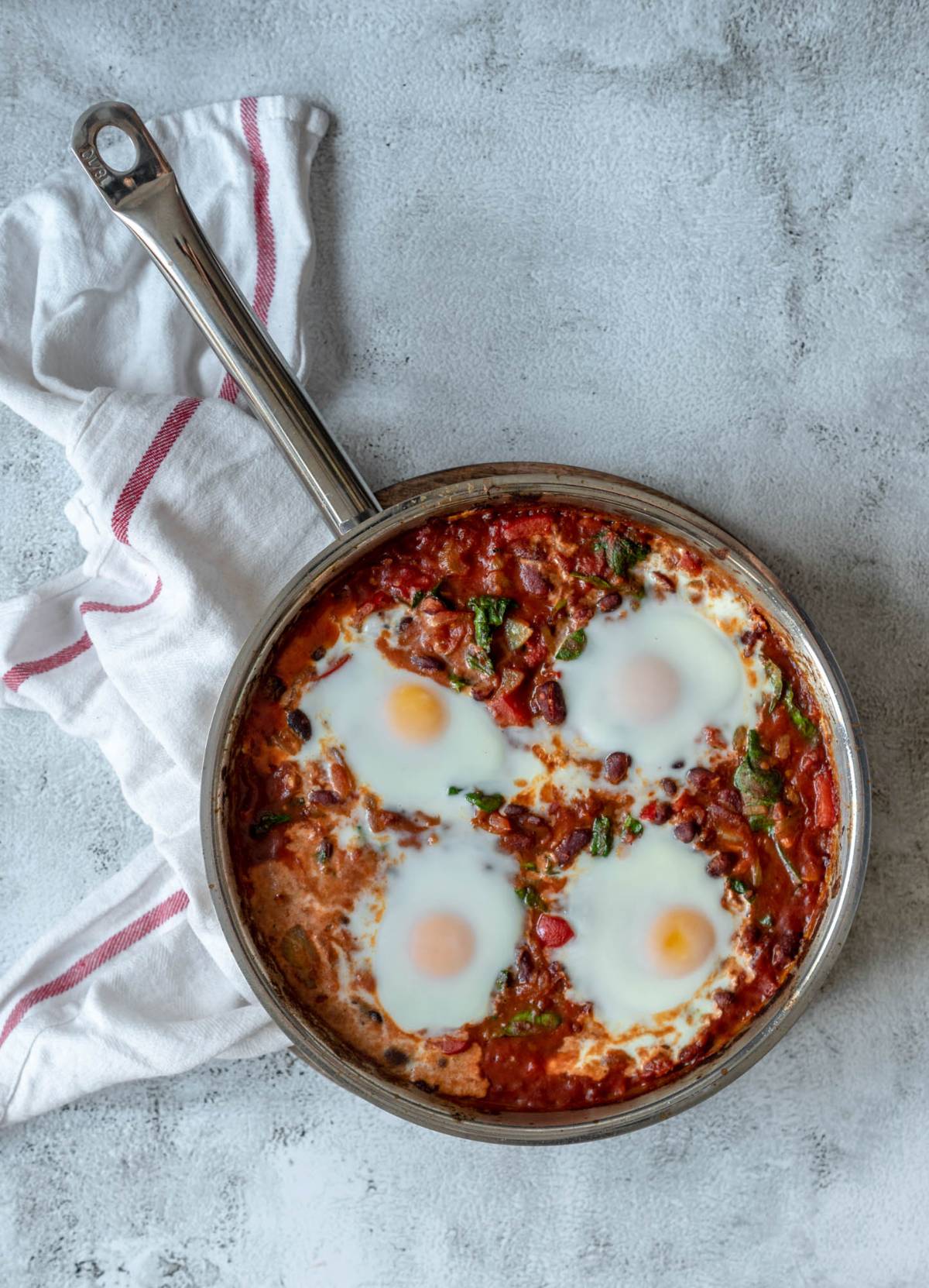 A skillet of spinach and red bean shakshuka with three poached eggs in tomato sauce, on a gray surface with a white towel—a perfect savoury breakfast recipe.