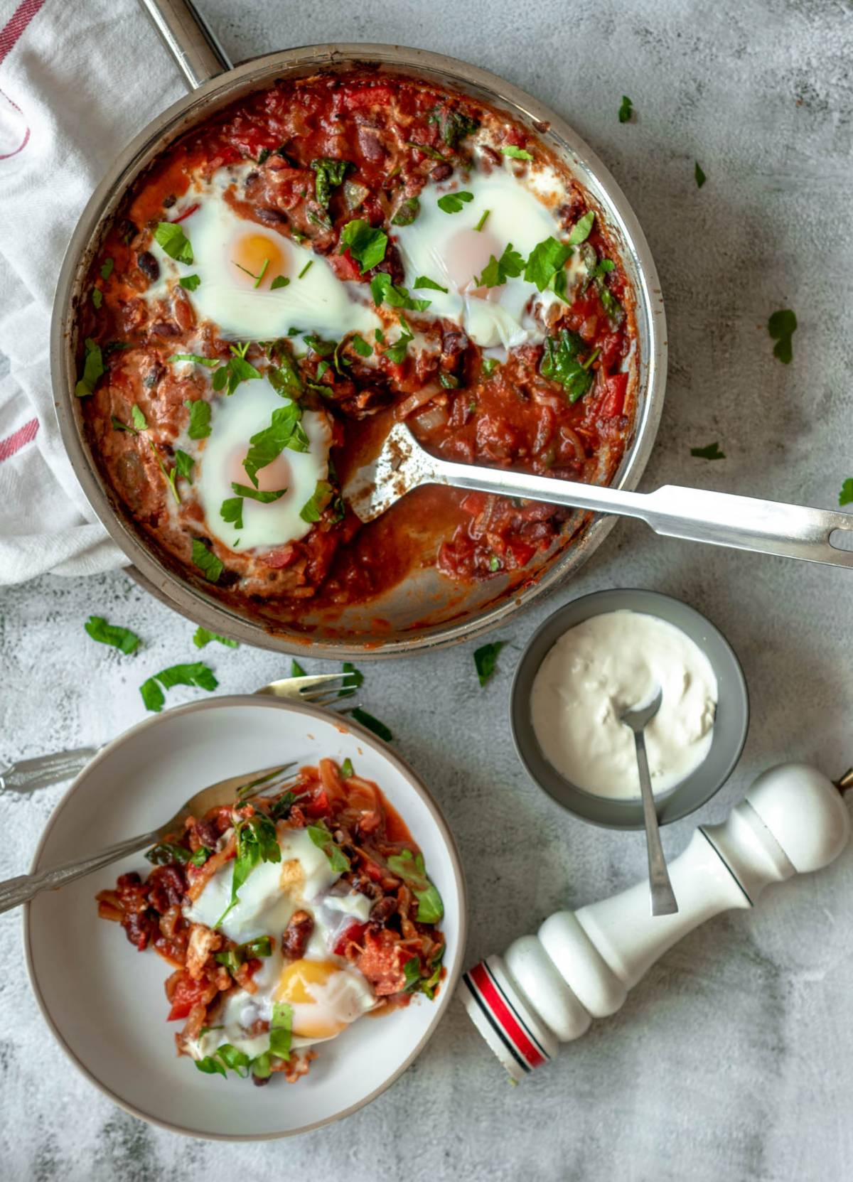 A pan of shakshuka with eggs poached in a tomato sauce, fresh herbs, and spinach, served with sour cream on a rustic table—this quick healthy meal is the perfect one pot meal for any time of day.