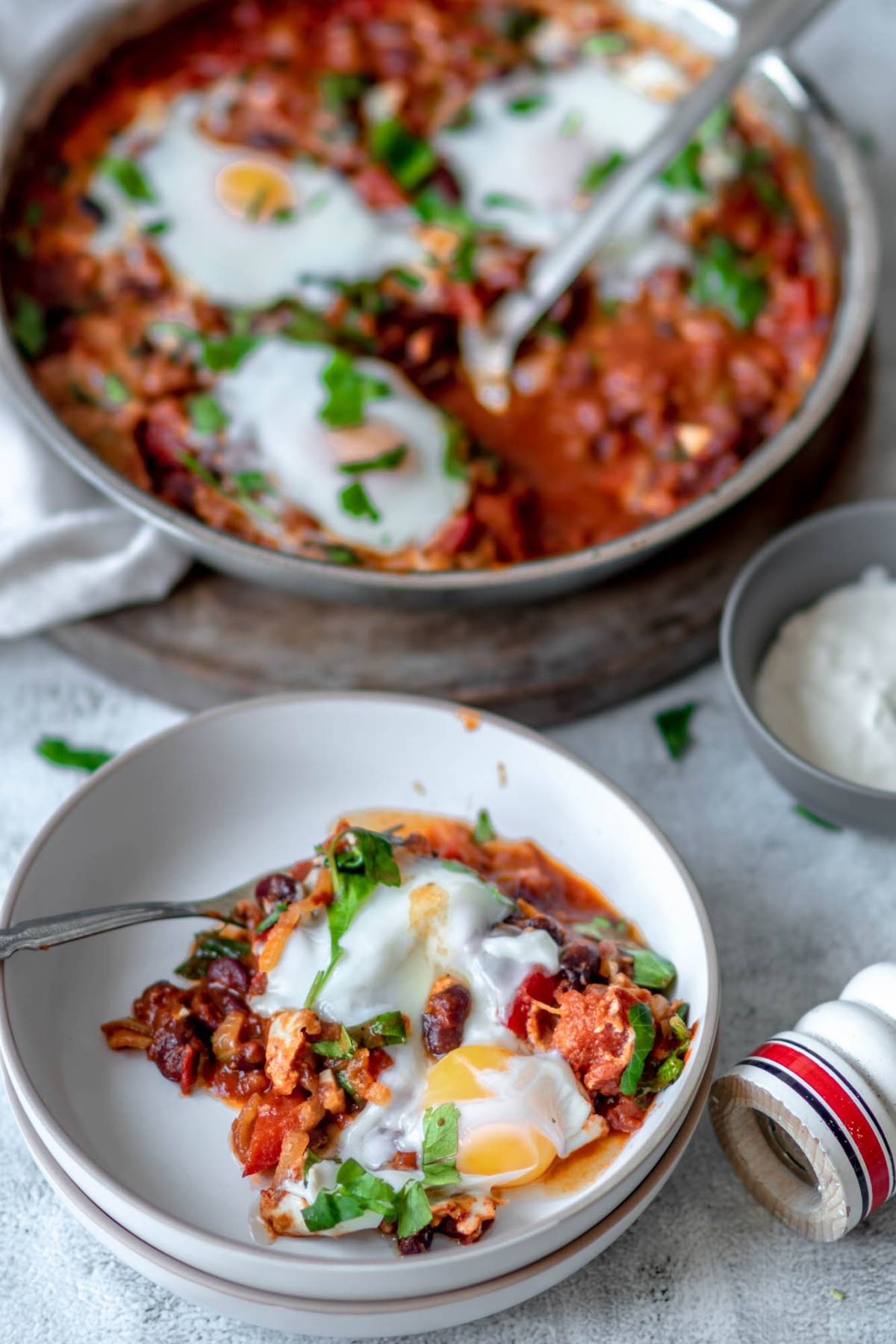 A plate of shakshuka with eggs poached in a tomato sauce and herbs, served next to a pan with more of this savoury breakfast recipe.