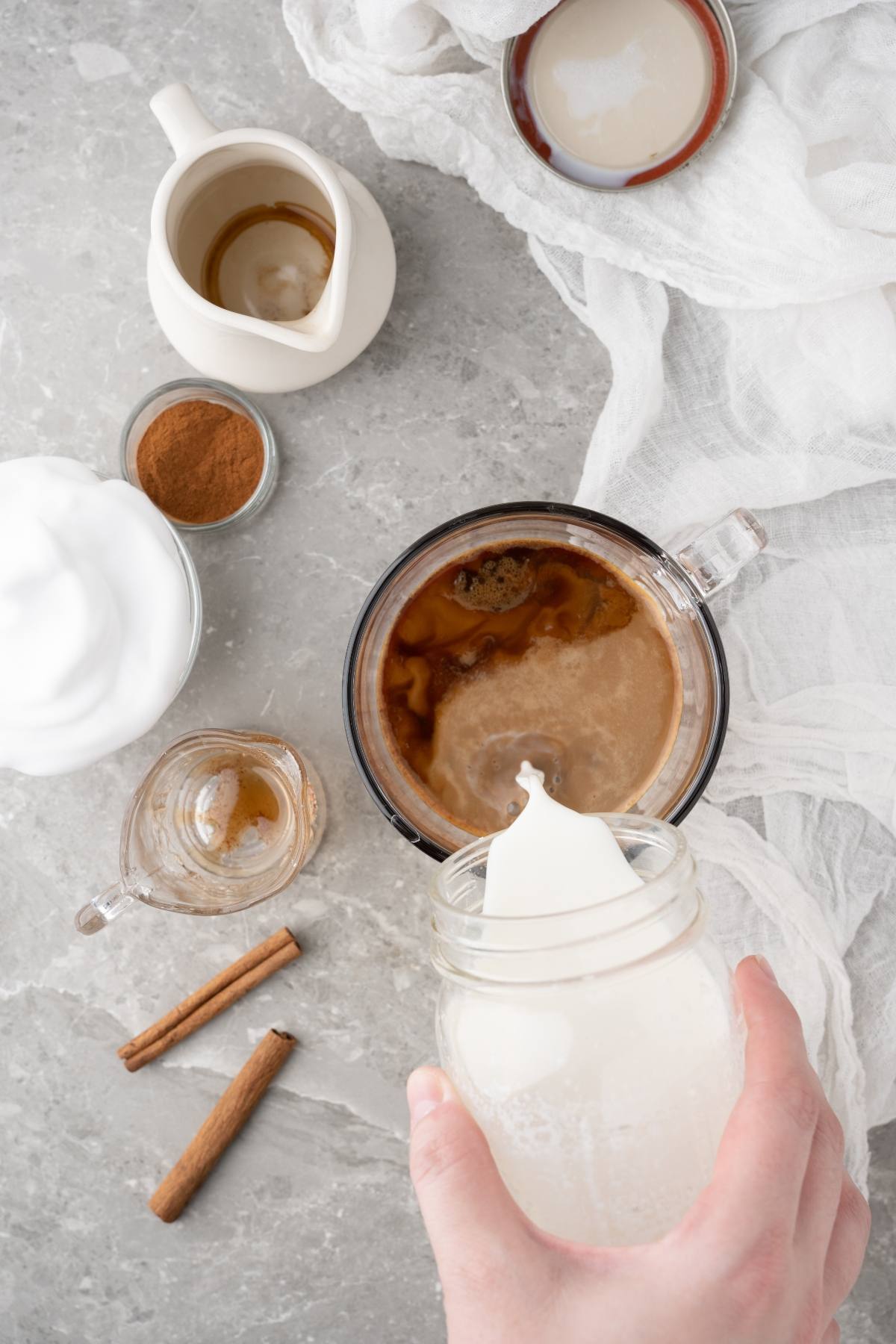 A hand pours milk into a mug of coffee, surrounded by cinnamon, whipped cream, and a jug on a gray surface—perfect for creating your own DIY cinnamon dolce latte at home.