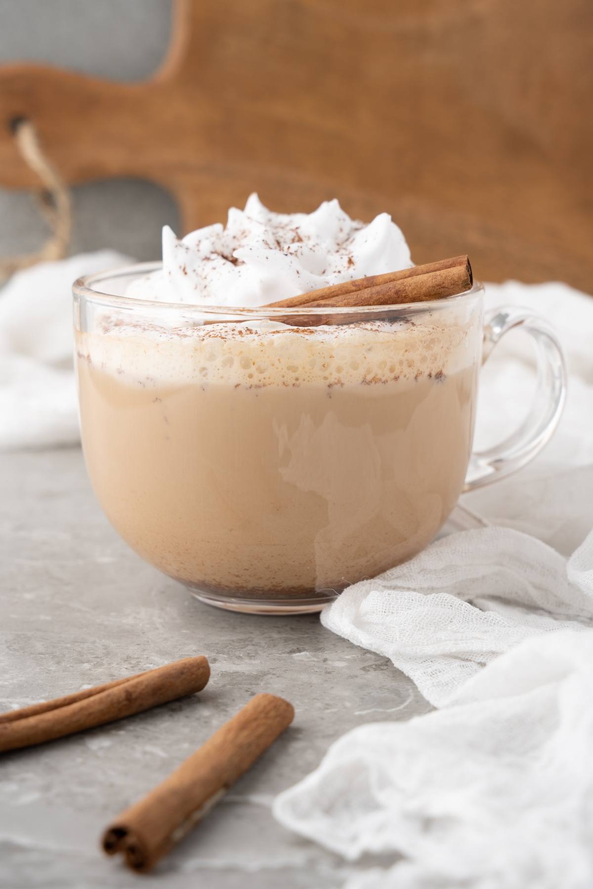 A glass mug of chai latte inspired by the Starbucks Cinnamon Dolce Latte, topped with whipped cream, cinnamon, and a cinnamon stick, on a gray surface.