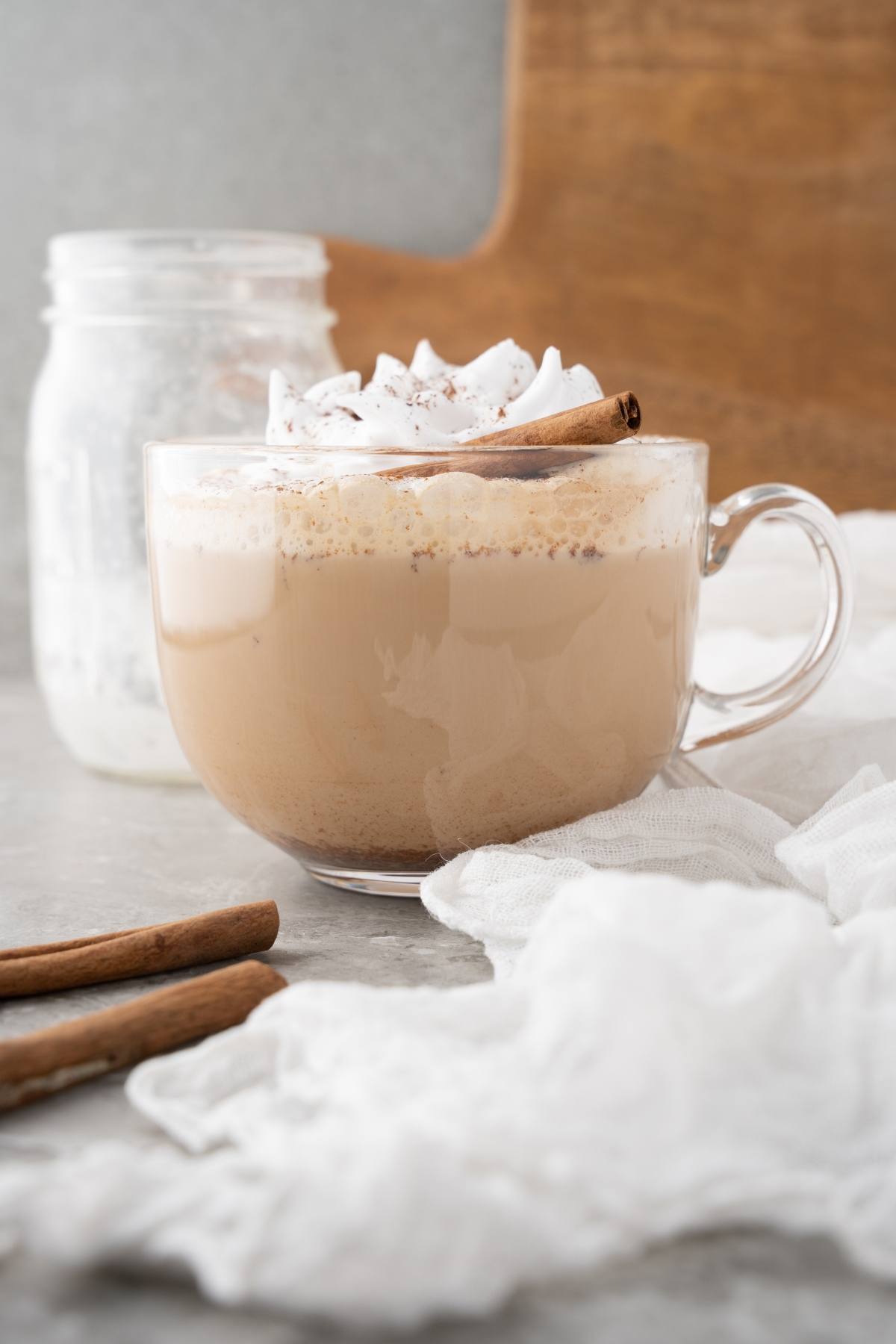 A glass mug of frothy latte topped with whipped cream and a cinnamon stick, with a jar of homemade cinnamon dolce syrup and cinnamon sticks nearby—a perfect Starbucks copycat latte.