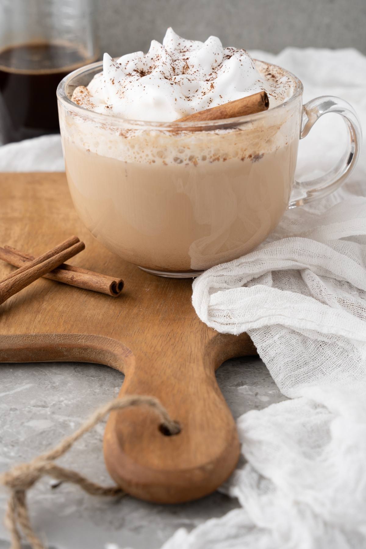 A glass mug of creamy coffee topped with whipped cream and a cinnamon stick sits on a wooden board—perfect for enjoying your DIY cinnamon dolce latte at home.