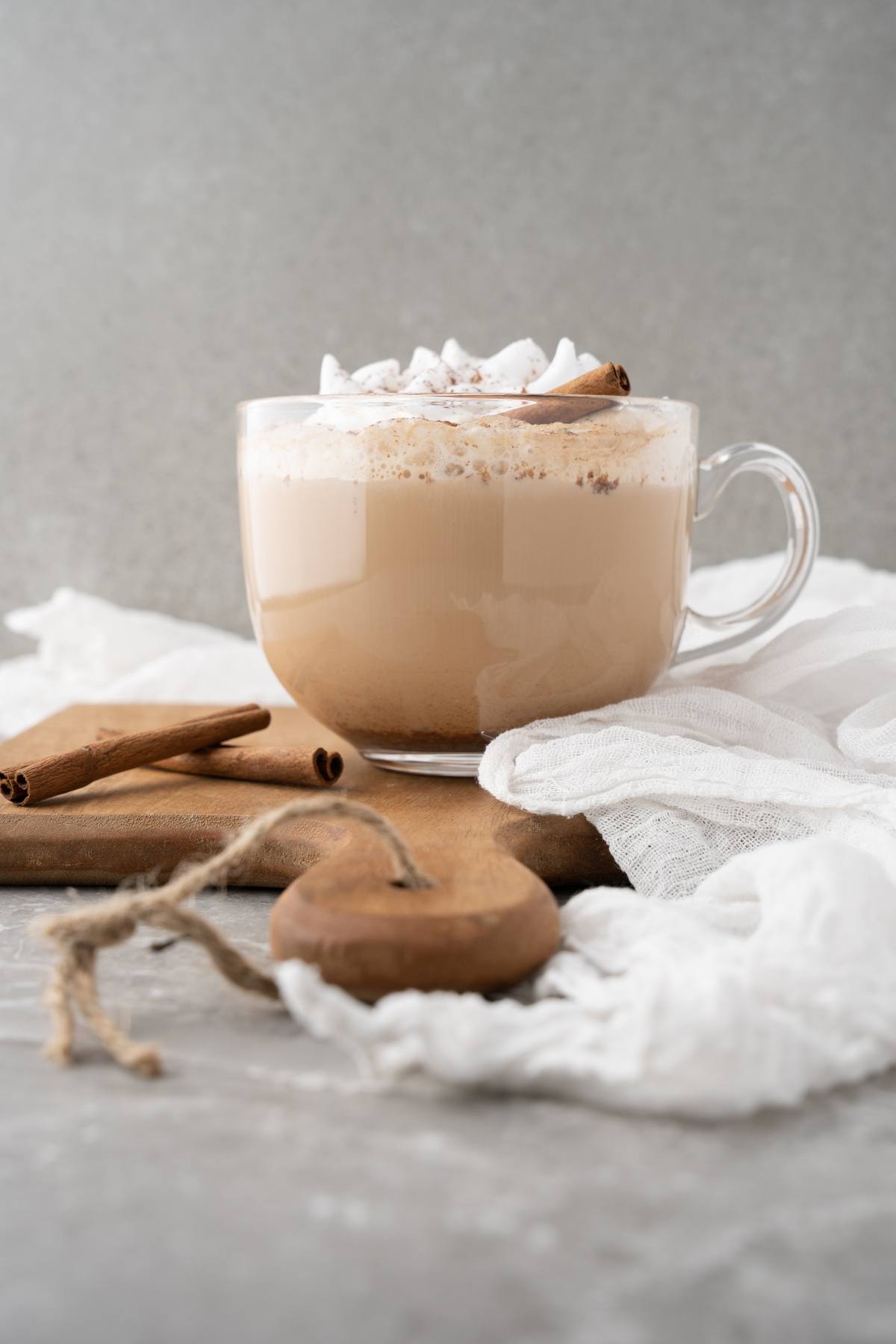 A glass mug of frothy latte, made with homemade cinnamon dolce syrup, topped with whipped cream and a cinnamon stick on a wooden board with white cloth.