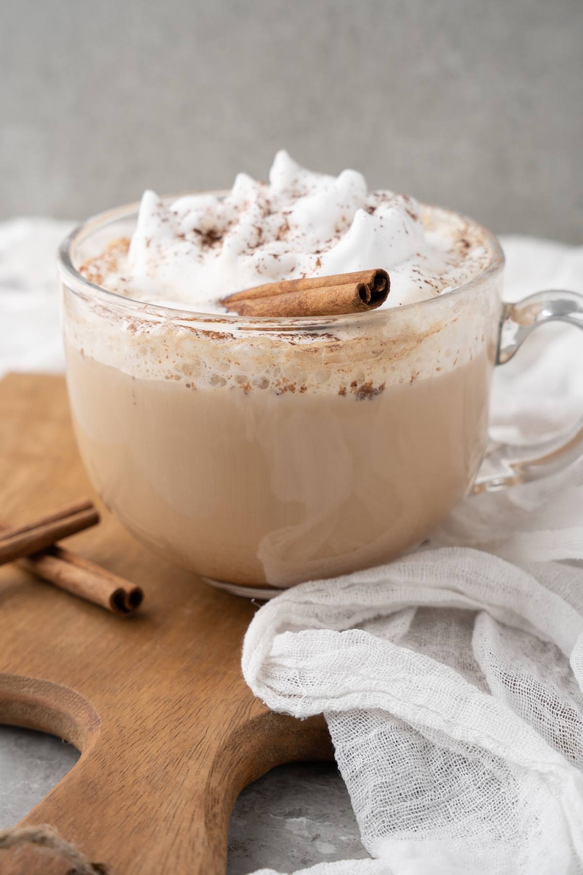 Clear glass mug of creamy coffee topped with whipped cream, cinnamon, and a cinnamon stick on a wooden board—just like a Starbucks Cinnamon Dolce Latte you can make at home.