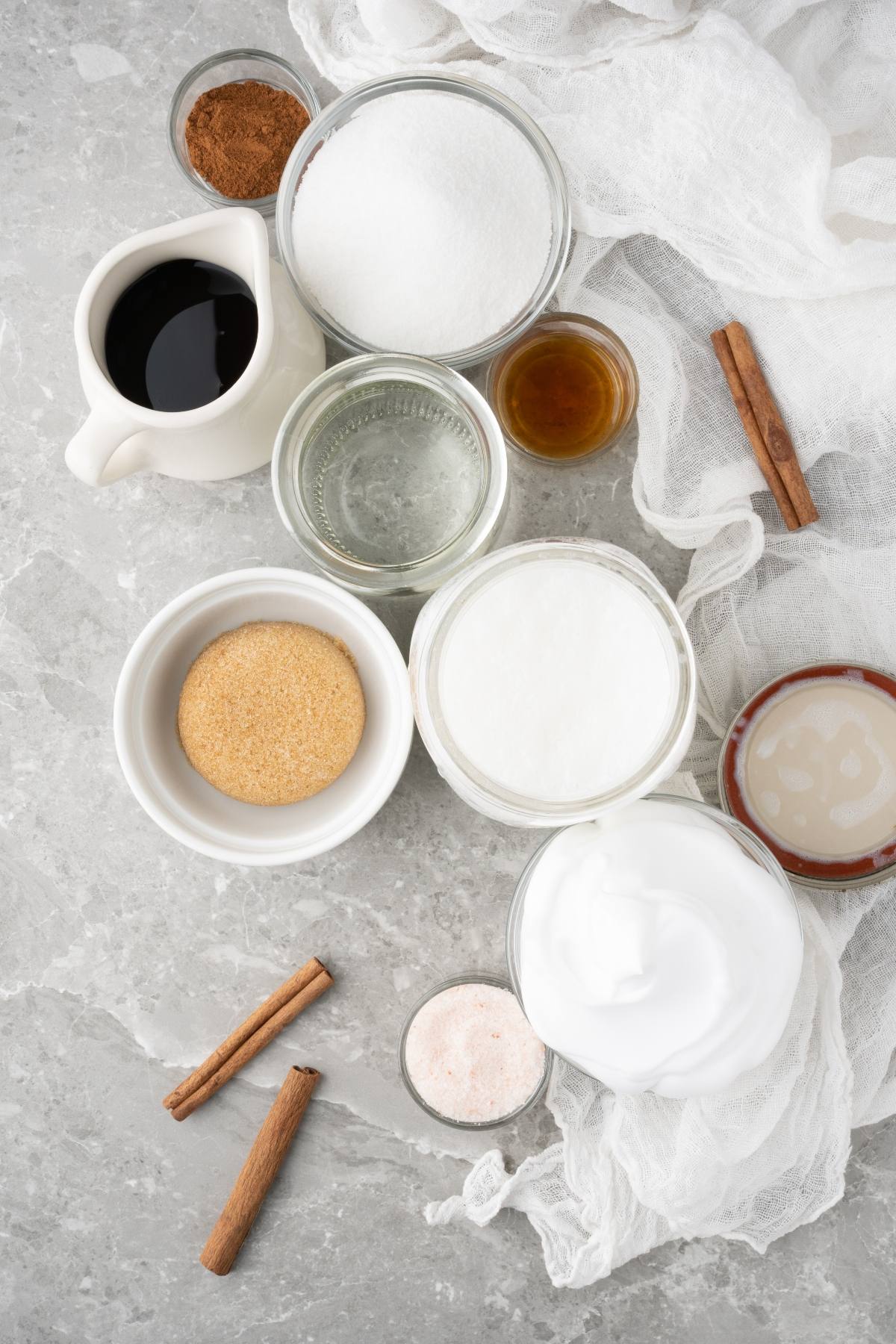 Assorted baking ingredients in bowls and jars on a gray surface with cinnamon sticks and white cloth—perfect for making homemade cinnamon dolce syrup to create a cozy dolce latte at home.