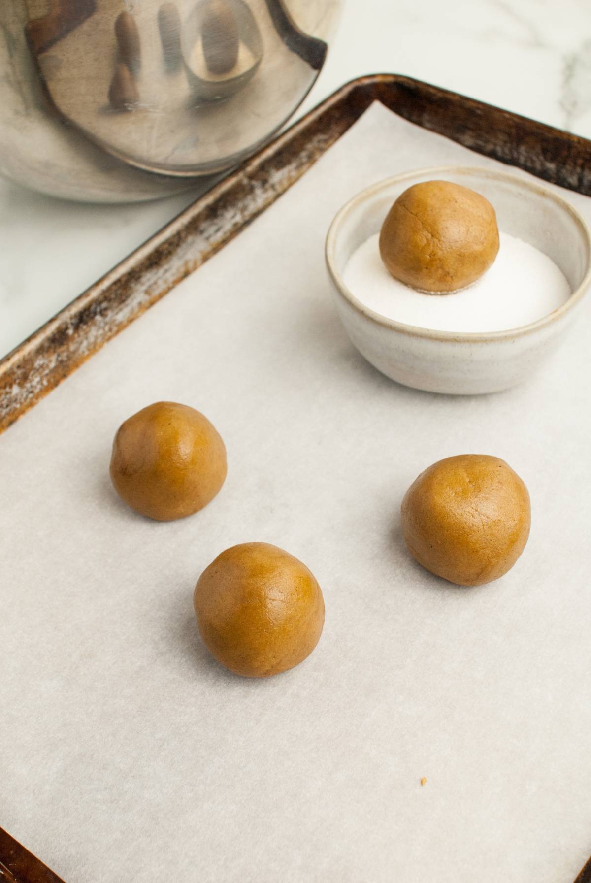 Four cookie dough balls on a parchment-lined baking sheet, with one ball in a small bowl of sugar—perfect for making Starbucks Copycat Ginger Molasses Cookies.