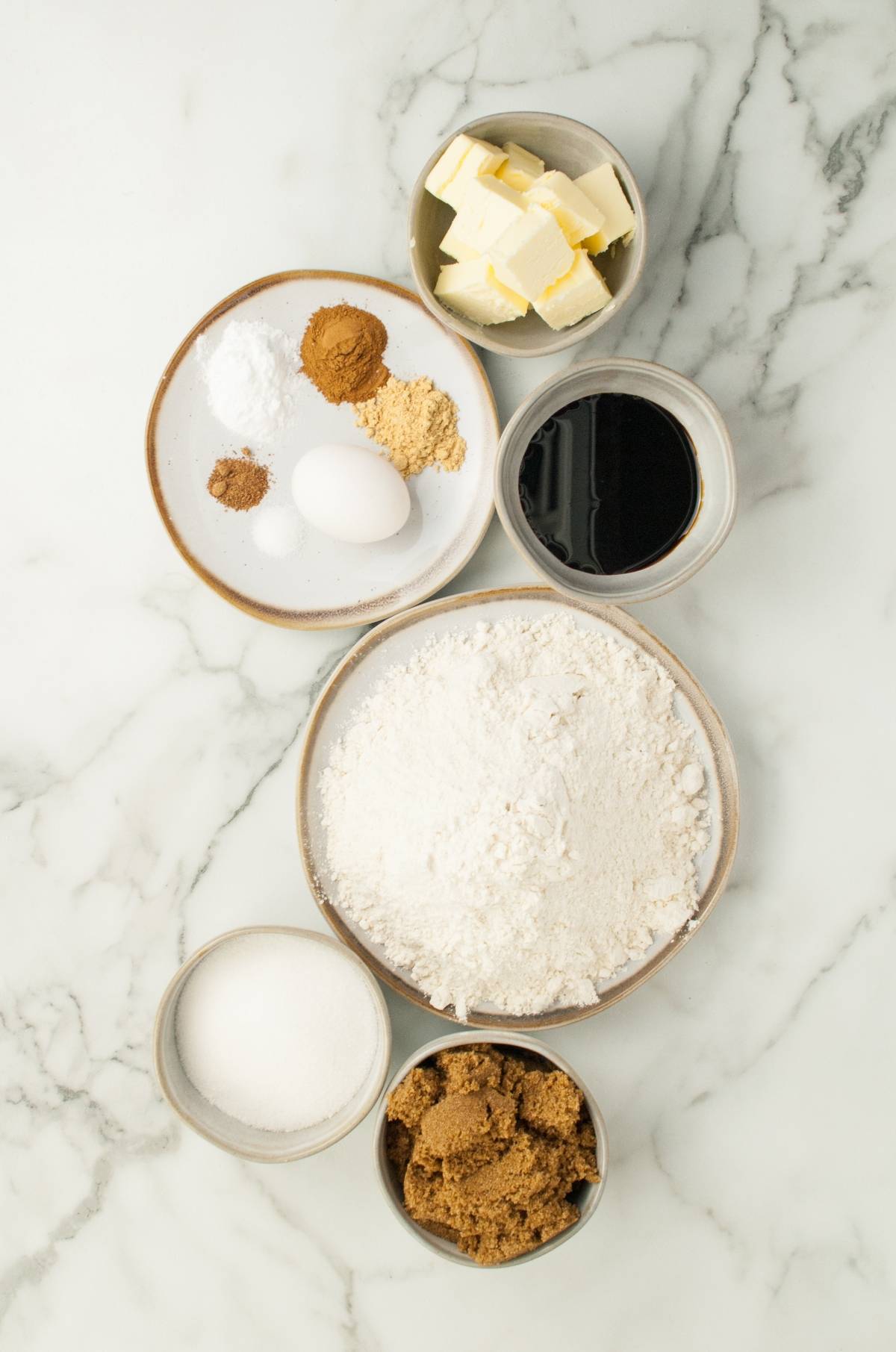 Bowls of flour, sugars, butter, molasses, spices, baking soda, and an egg on a marble surface—everything you need to make Starbucks Copycat Ginger Molasses Cookies at home.