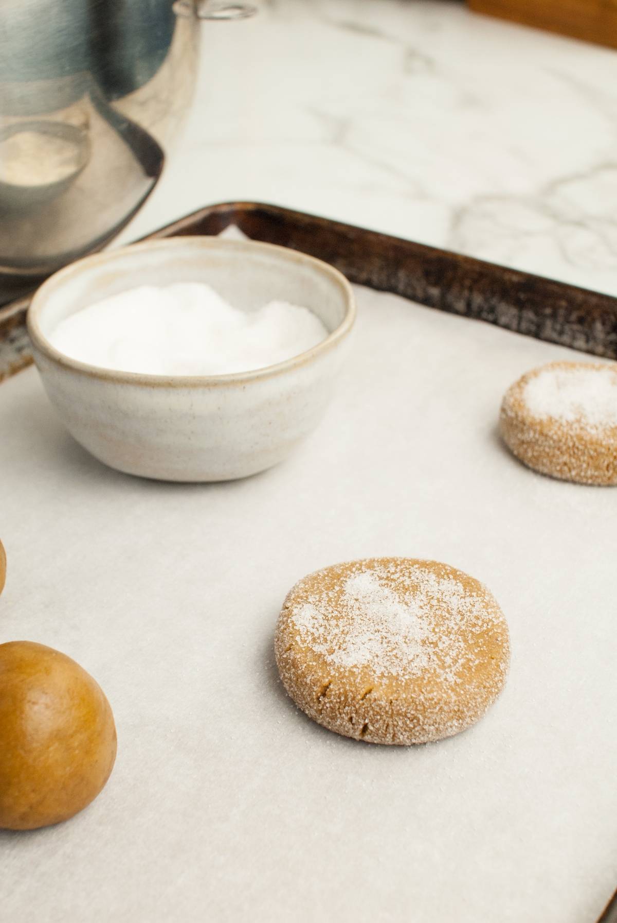 A baking tray with cookie dough balls and a sugared cookie next to a bowl of sugar on parchment paper, perfect for making Homemade Starbucks Christmas cookies or delicious Starbucks Copycat Ginger Molasses Cookies.