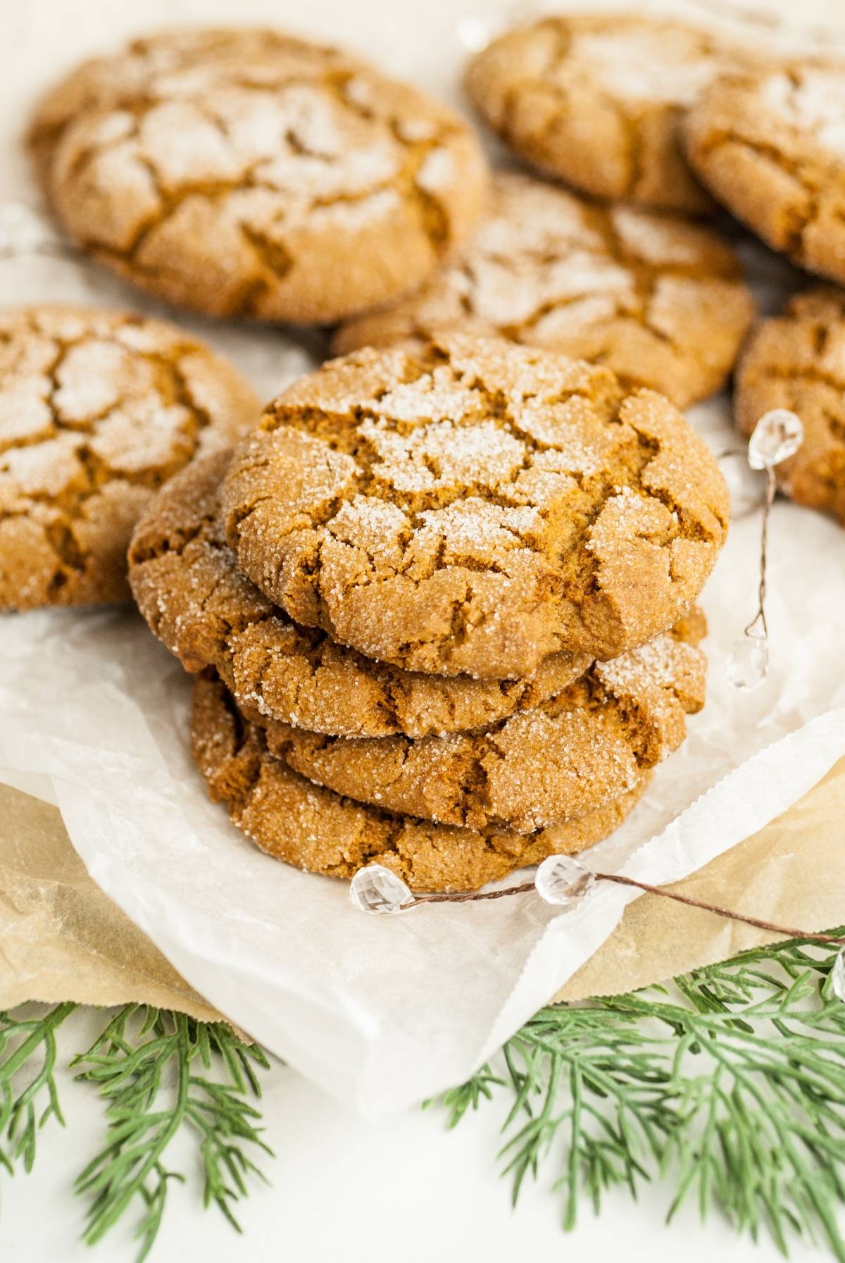 A stack of sugar-coated gingersnap molasses cookies on parchment paper with festive greenery nearby, reminiscent of the best ginger molasses cookie recipe.