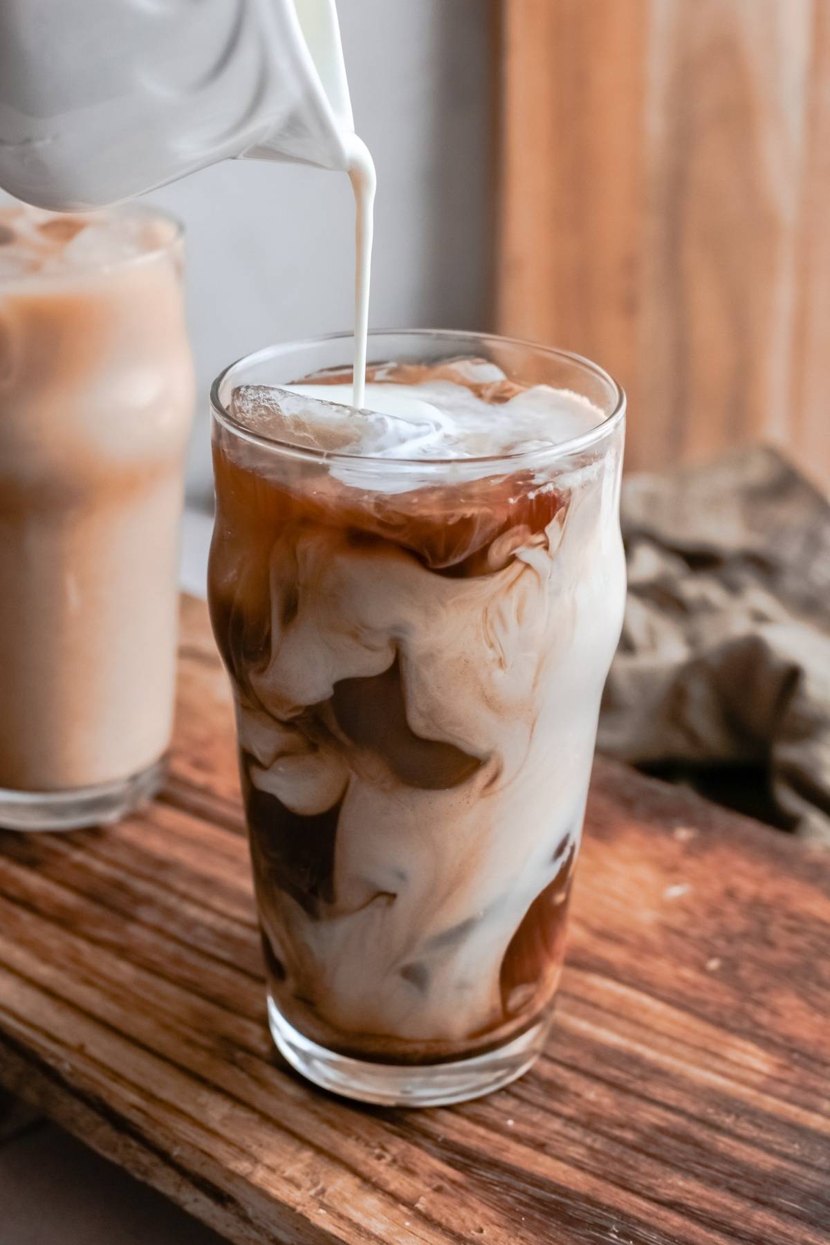 Milk being poured into a glass of iced coffee, creating beautiful swirls—just like making your own DIY Starbucks shaken espresso—with another drink in the background.
