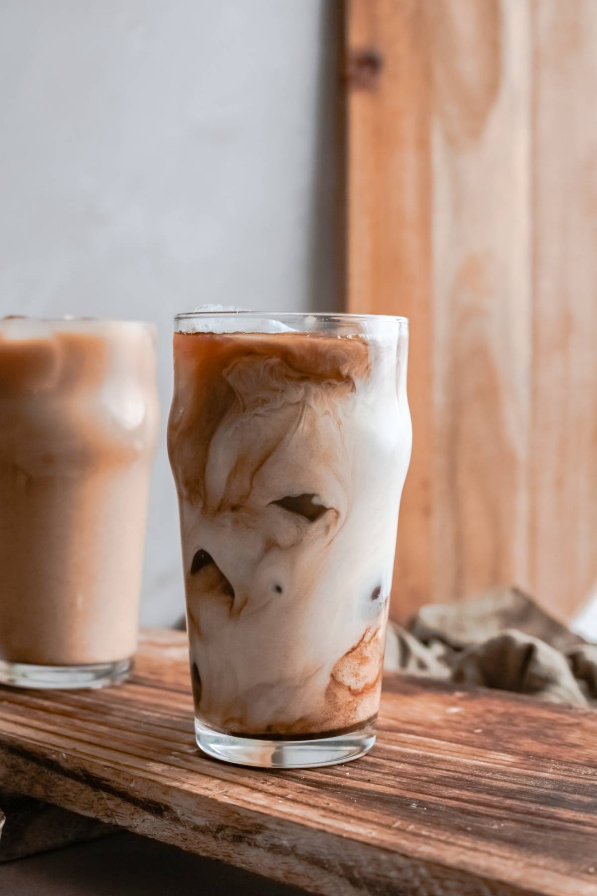 A glass of iced coffee with swirling cream on a wooden board, just like a copycat Starbucks iced drink—perfect for recreating your favorite café moments at home. Another coffee glass rests in the background.