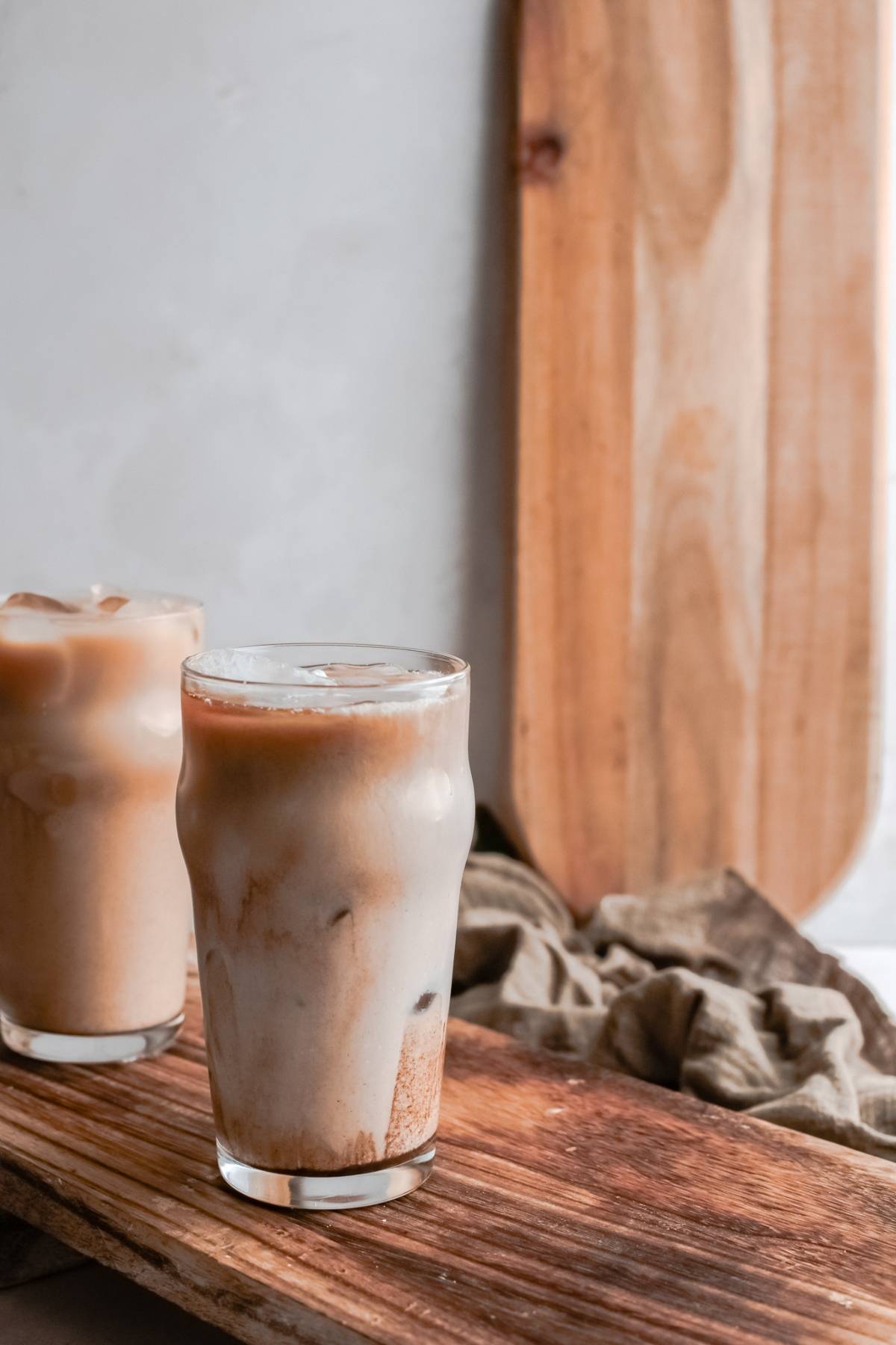 Two glasses of iced coffee with milk on a wooden tray, perfect for a DIY Starbucks shaken espresso vibe, with a wooden board and cloth in the background.