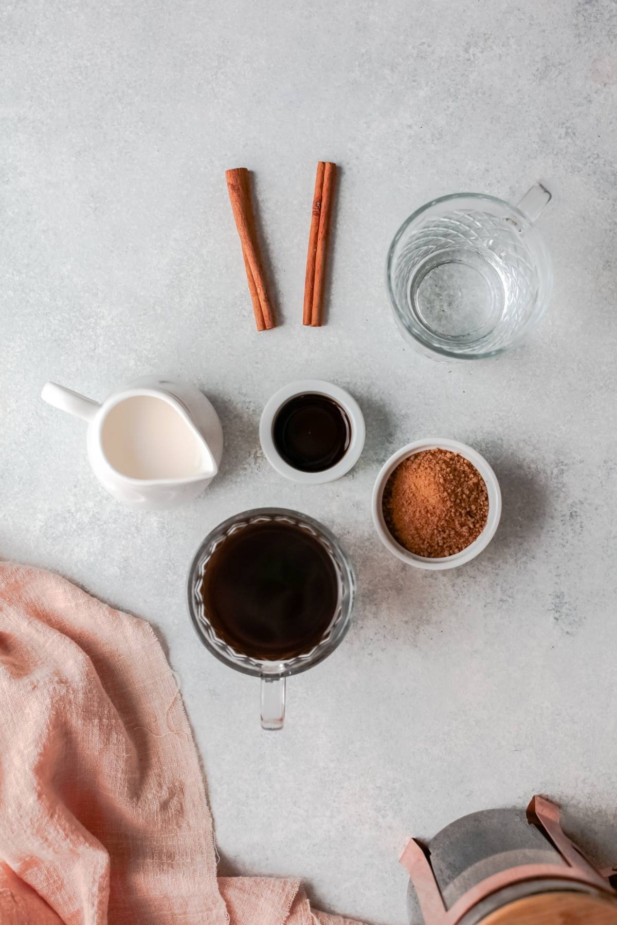 Coffee, milk, vanilla, brown sugar, water, and two cinnamon sticks arranged on a light surface with a cloth—perfect for DIY Starbucks shaken espresso or to make a Starbucks iced drink at home.