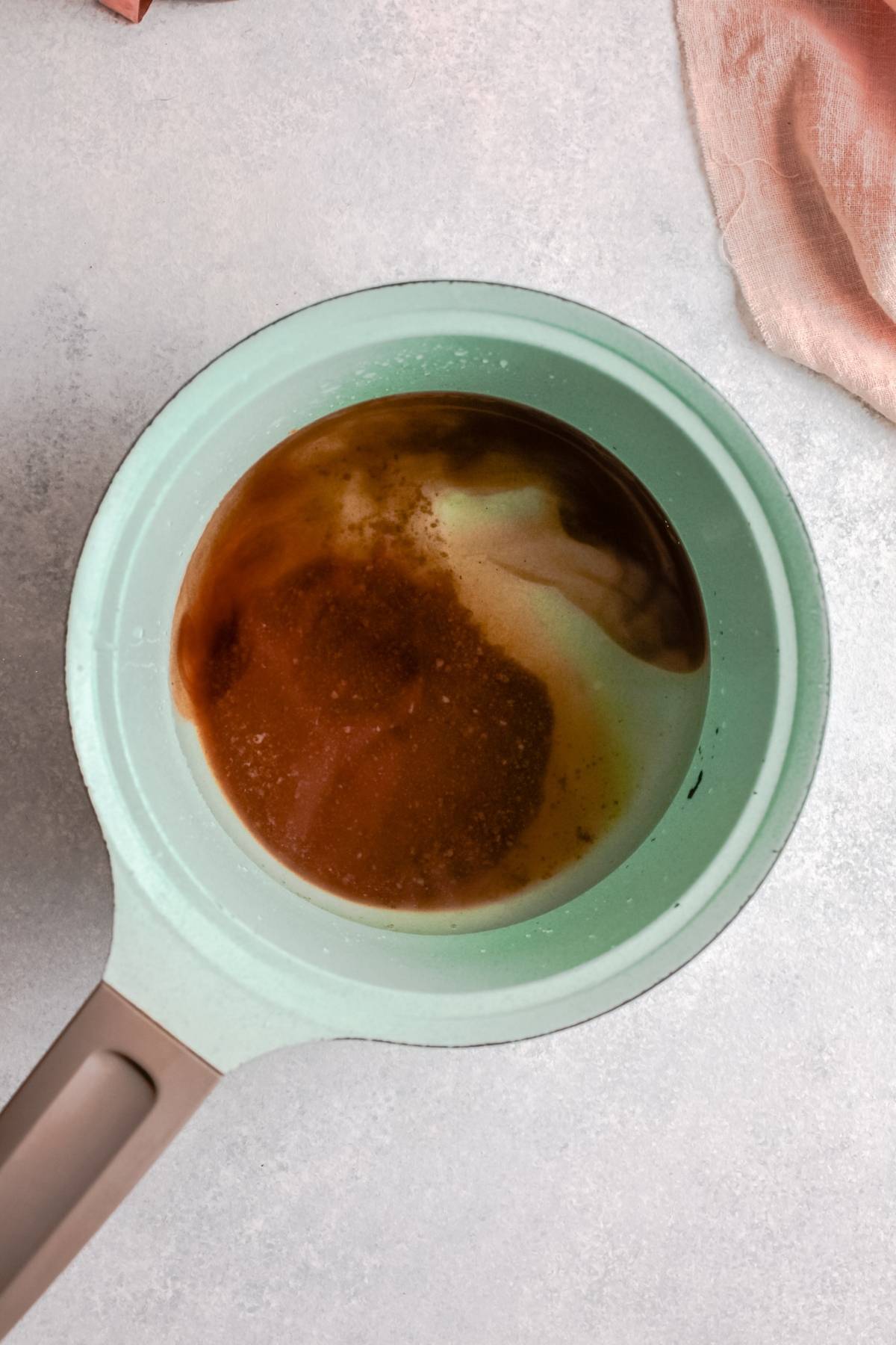 A light green saucepan with a brown and white liquid mixture, perfect for making Homemade brown sugar syrup for your Starbucks Copycat Iced Brown Sugar Oatmilk Shaken Espresso, sits on a light countertop near a pink cloth.