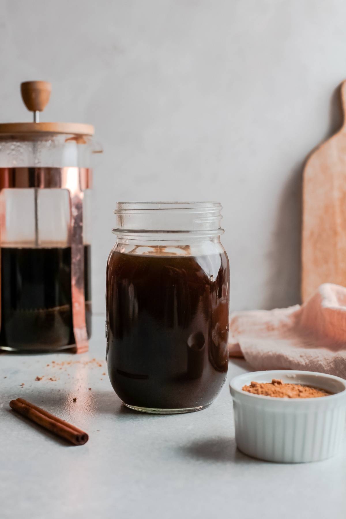 A jar of iced coffee sits on a table with a French press, cinnamon stick, and a bowl of powder—perfect for making a Starbucks Copycat Iced Brown Sugar Oatmilk Shaken Espresso at home.