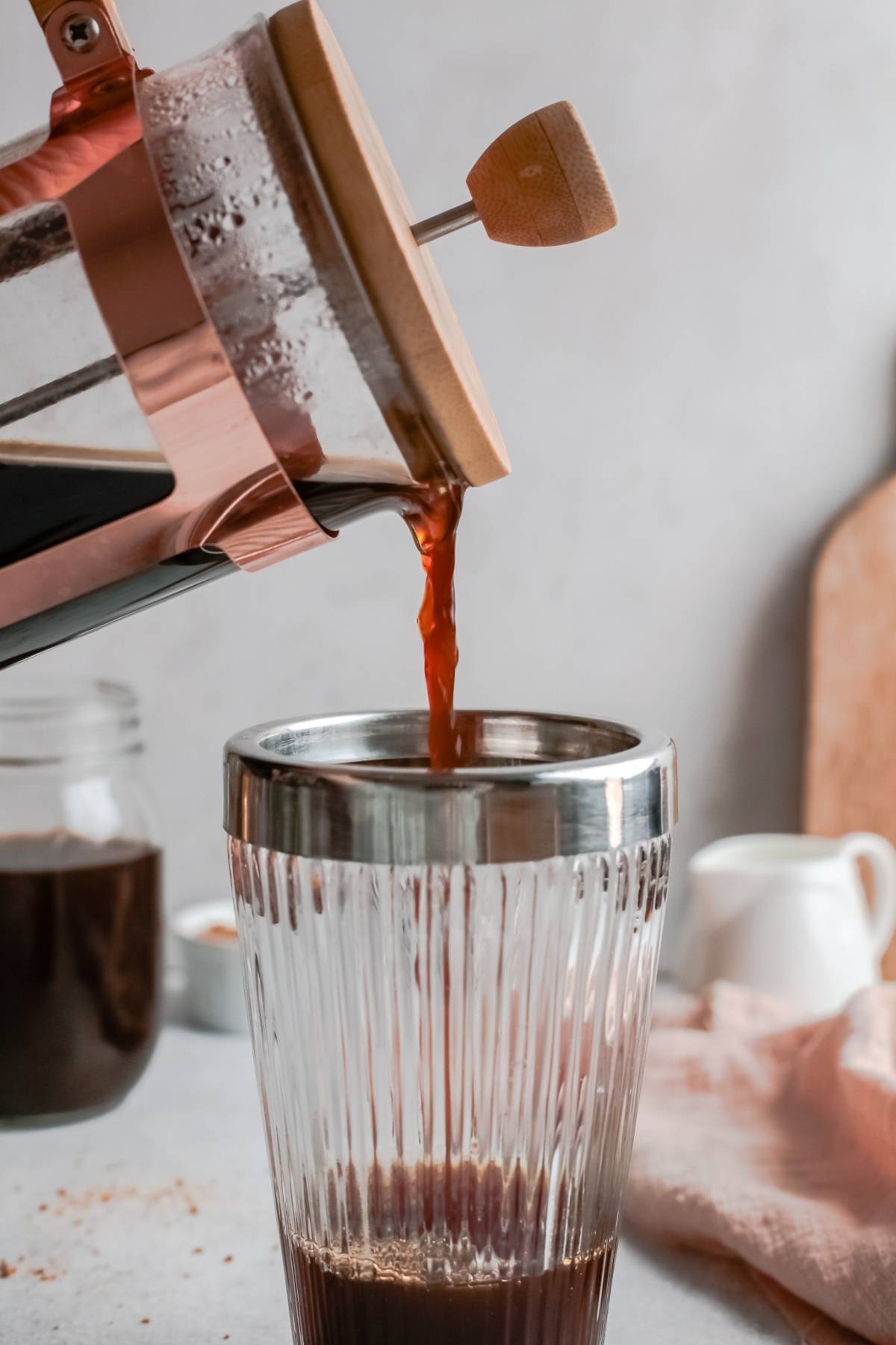 Coffee being poured from a French press into a clear glass with a metal rim on a kitchen counter, perfect for making your own DIY Starbucks shaken espresso with homemade brown sugar syrup.