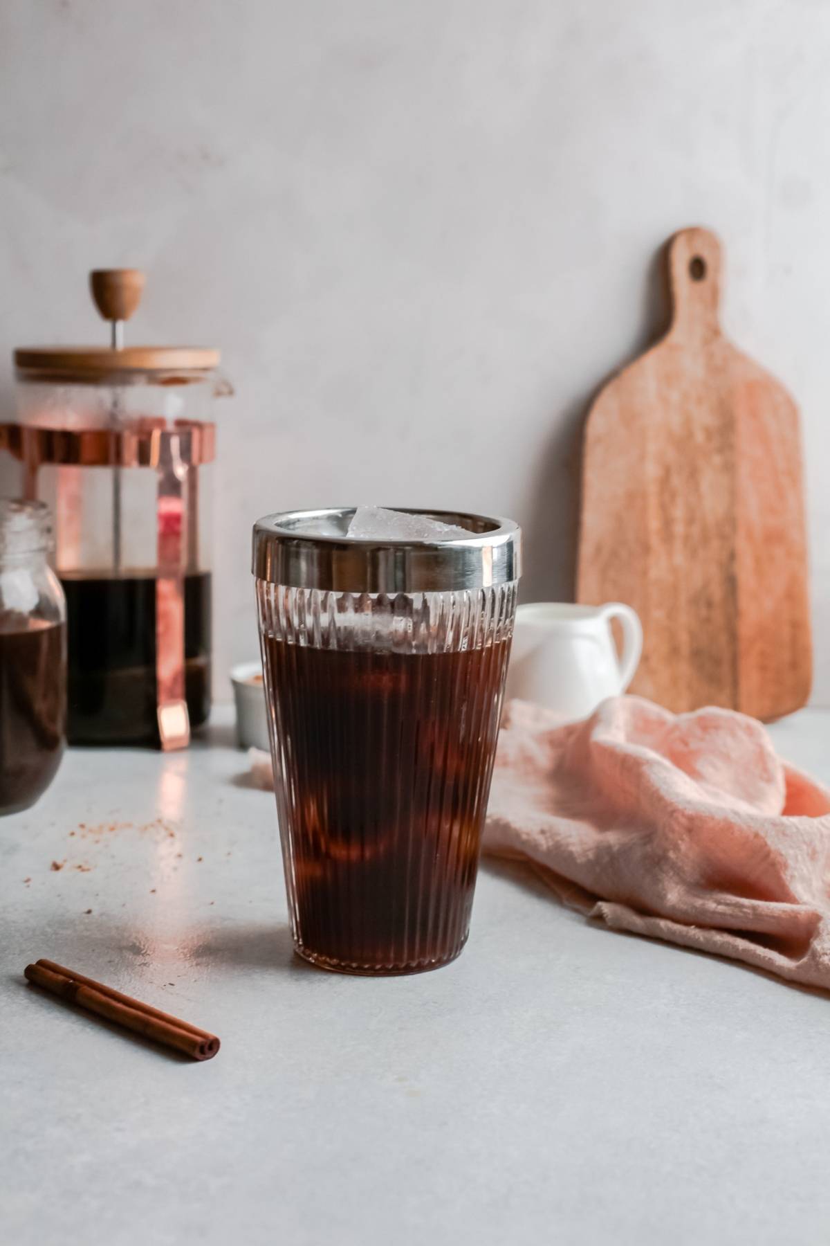 A glass of iced coffee sits on a counter near a French press, cutting board, and cloth napkin—perfect for your DIY Starbucks shaken espresso at home.