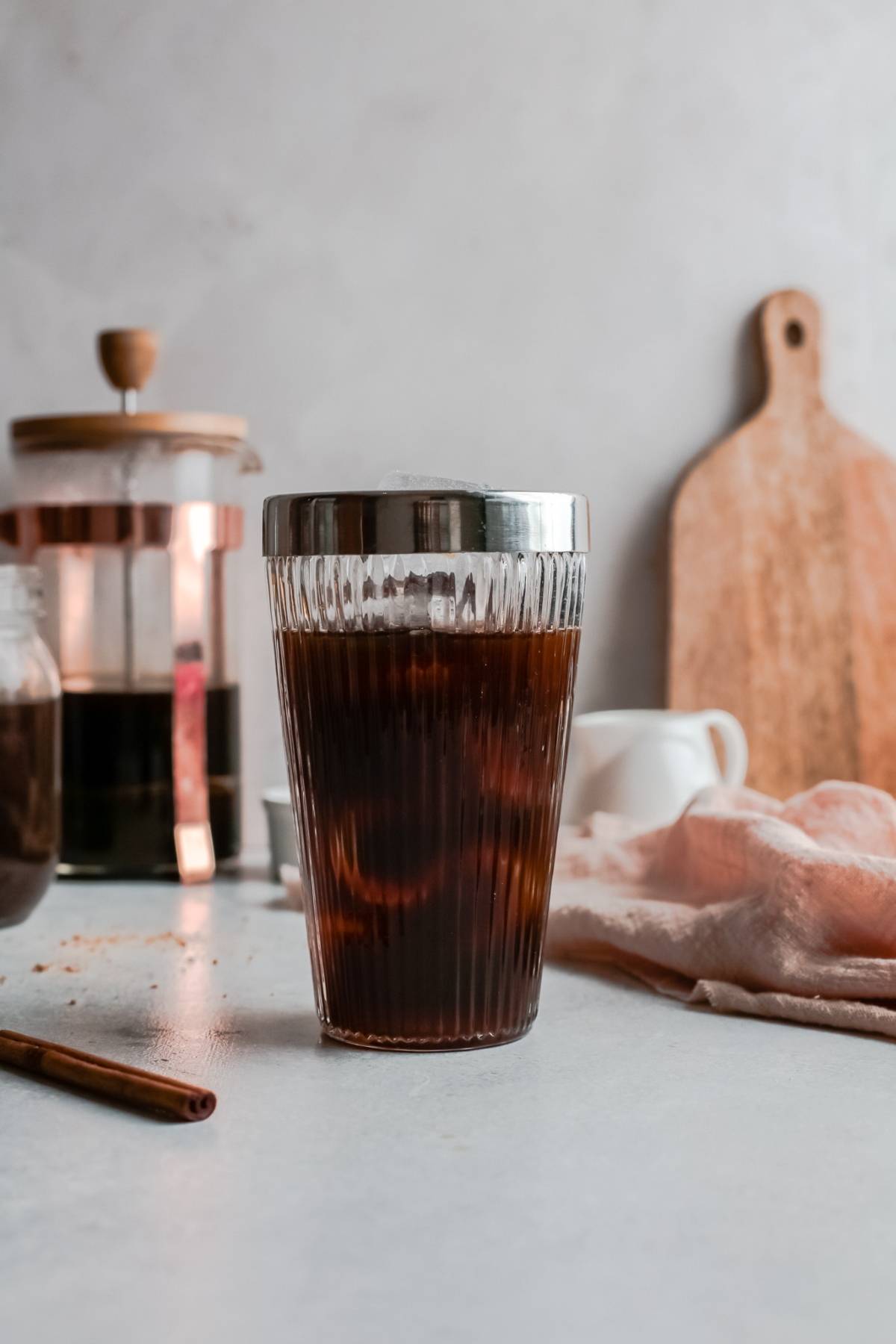 A glass of iced coffee sits on a table with a French press, cutting board, and napkin—perfect for making a Starbucks Copycat Iced Brown Sugar Oatmilk Shaken Espresso at home using homemade brown sugar syrup.