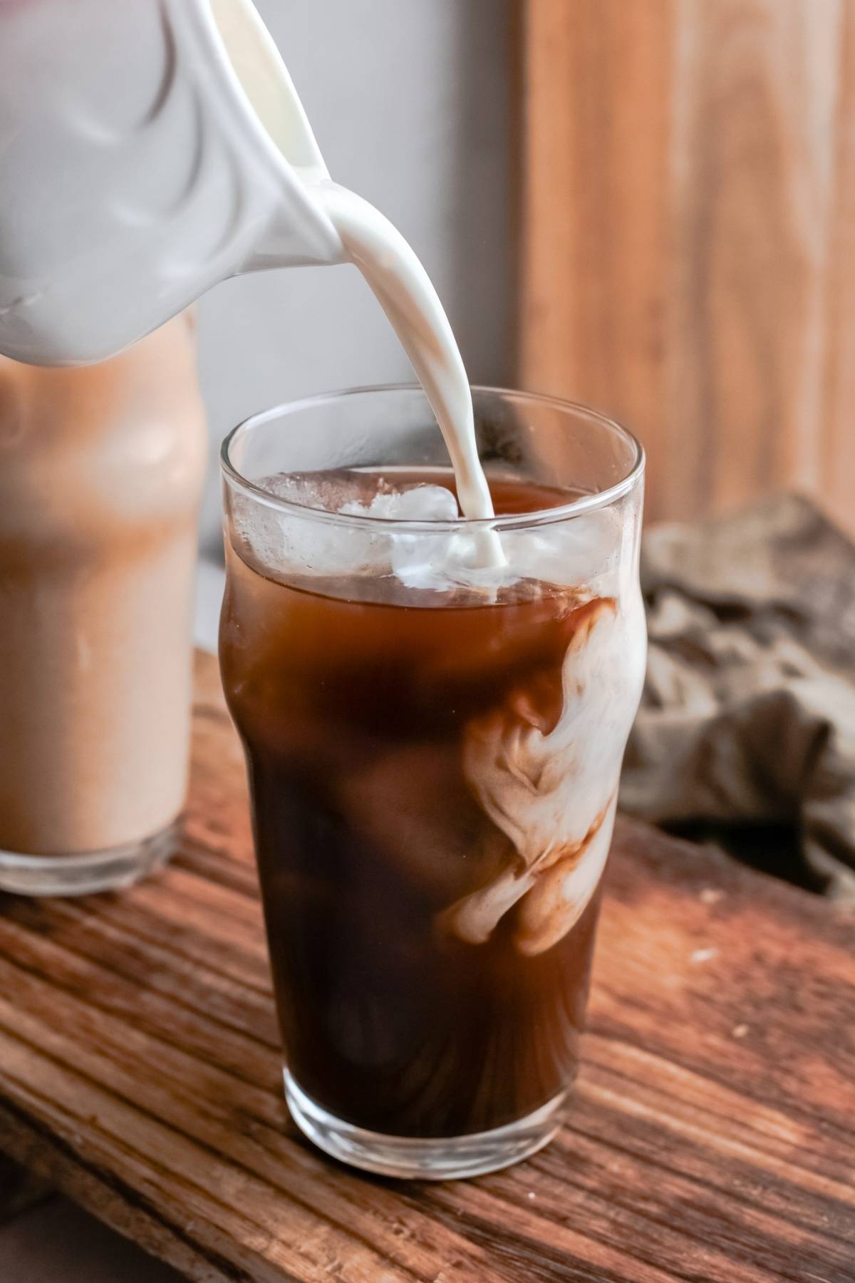 Milk being poured into a glass of iced coffee on a wooden surface, creating creamy swirls—just like making a Starbucks Copycat Iced Brown Sugar Oatmilk Shaken Espresso at home.
