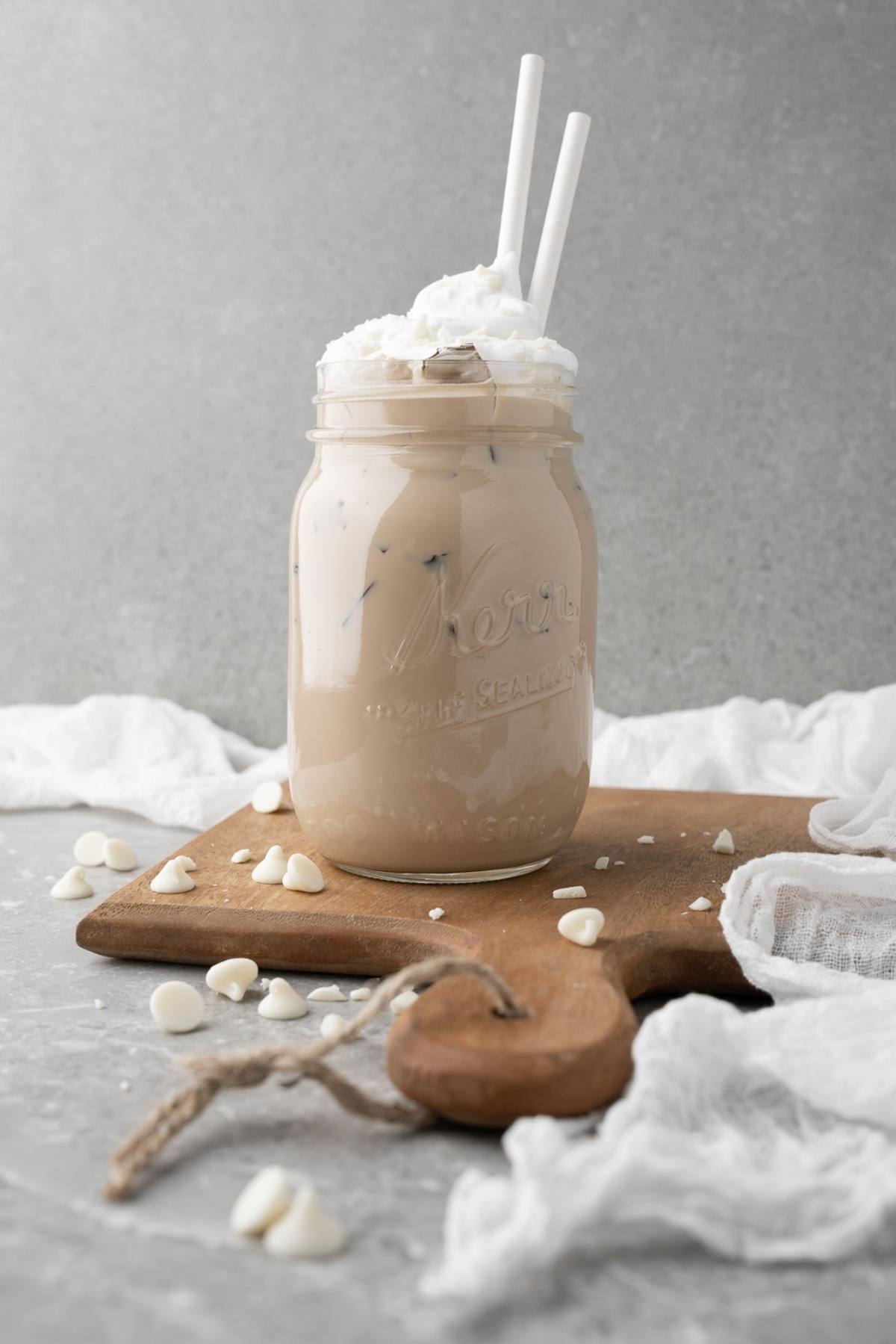 Iced coffee with whipped cream in a mason jar, two straws, on a wooden board with white chocolate chips—a true Starbucks Copycat Iced White Chocolate Mocha you can make at home.